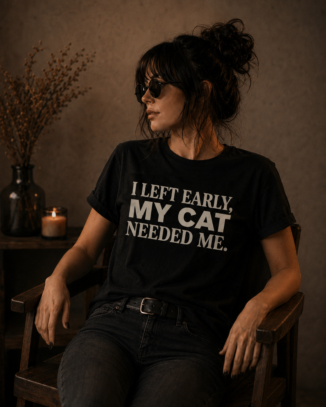 A woman wearing a black graphic t-shirt with the text 'I LEFT EARLY, MY CAT NEEDED ME.' in white, sitting in a rustic wooden chair in a dimly lit room.