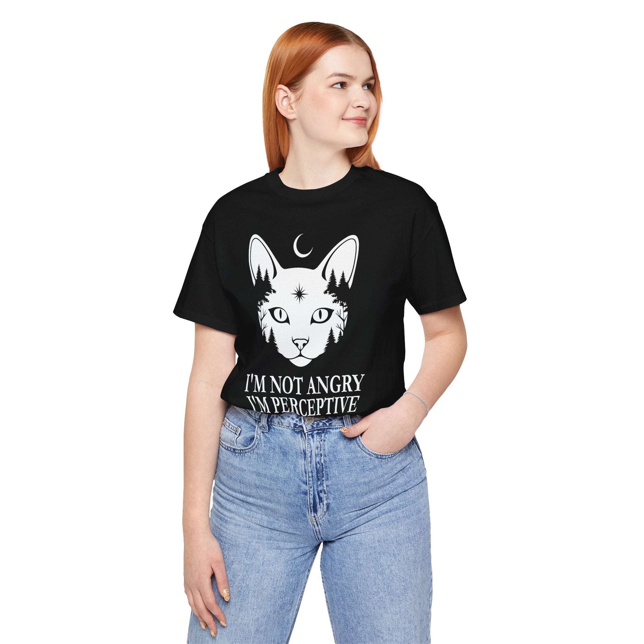 A woman with red hair wearing a black graphic t-shirt with a white cat illustration and the text 'I'M NOT ANGRY I'M PERCEPTIVE', paired with light blue jeans, against a plain white background.