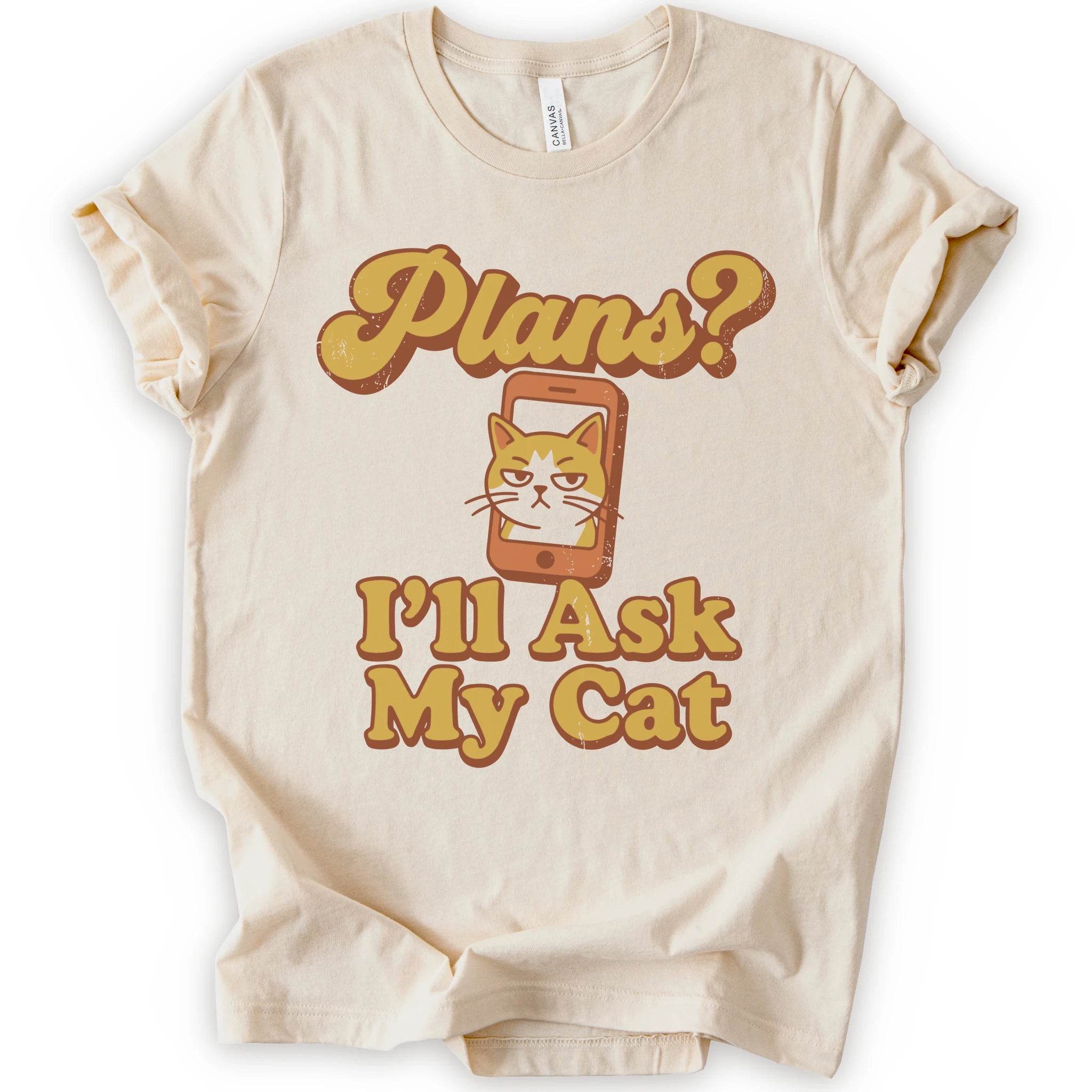 A natural crewneck t-shirt with a graphic print that reads 'Plans? I'll Ask My Cat' in a retro yellow-orange font, featuring a grumpy orange tabby cat cartoon inside a smartphone.