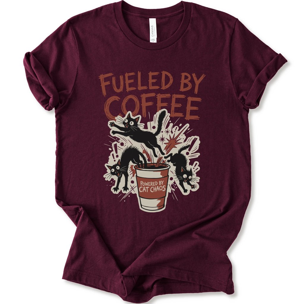 A maroon short-sleeve graphic t-shirt laid flat on a white background. The shirt features a distressed graphic print with the text 'FUELED BY COFFEE' in a reddish-brown color, above an illustration of black cats emerging from a coffee cup labeled 'POWERED BY CAT CHAOS'.