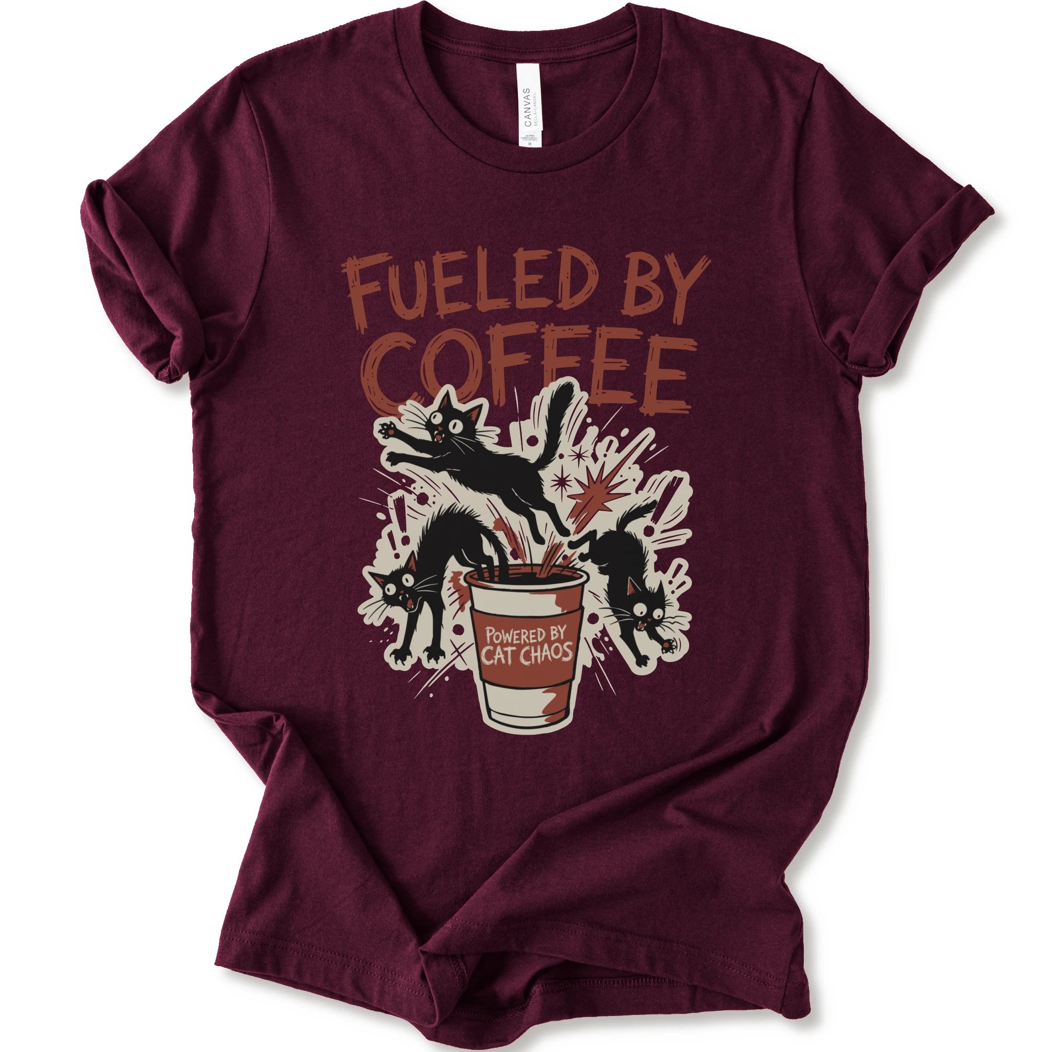 A maroon short-sleeve graphic t-shirt laid flat on a white background. The shirt features a distressed graphic print with the text 'FUELED BY COFFEE' in a reddish-brown color, above an illustration of black cats emerging from a coffee cup labeled 'POWERED BY CAT CHAOS'.