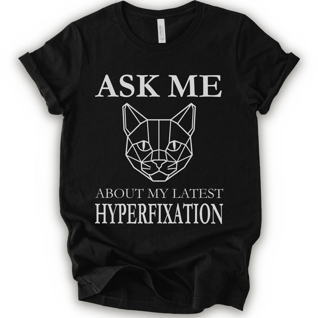 A black short-sleeve t-shirt with a white graphic print featuring a geometric cat face and the text 'ASK ME ABOUT MY LATEST HYPERFIXATION'.