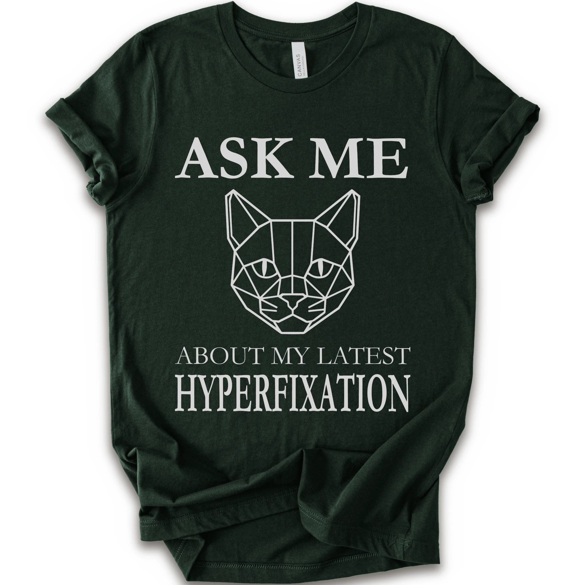 A forest short-sleeve t-shirt with a white graphic print featuring a geometric cat face and the text 'ASK ME ABOUT MY LATEST HYPERFIXATION'.