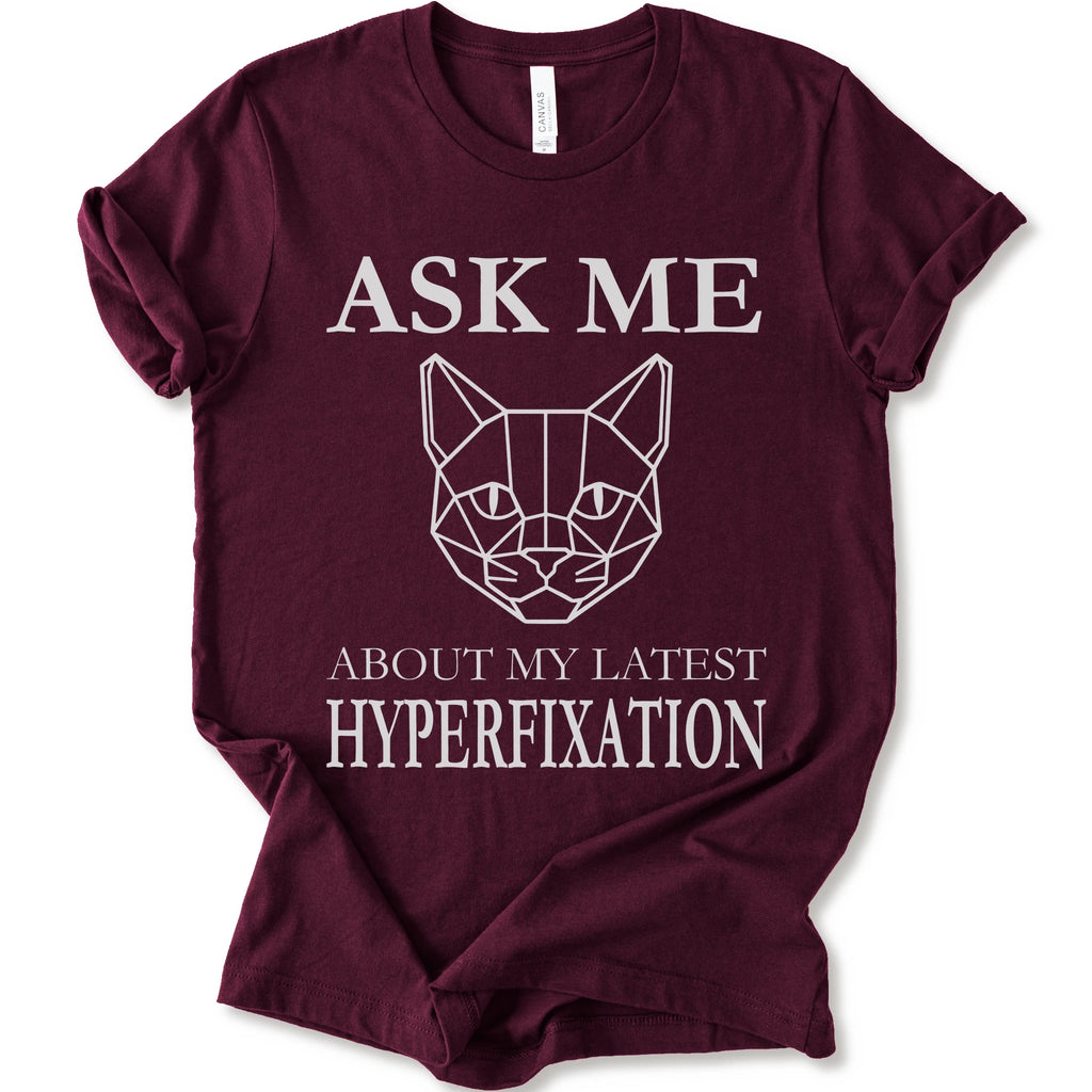 A maroon short-sleeve t-shirt with a white graphic print featuring a geometric cat face and the text 'ASK ME ABOUT MY LATEST HYPERFIXATION'.