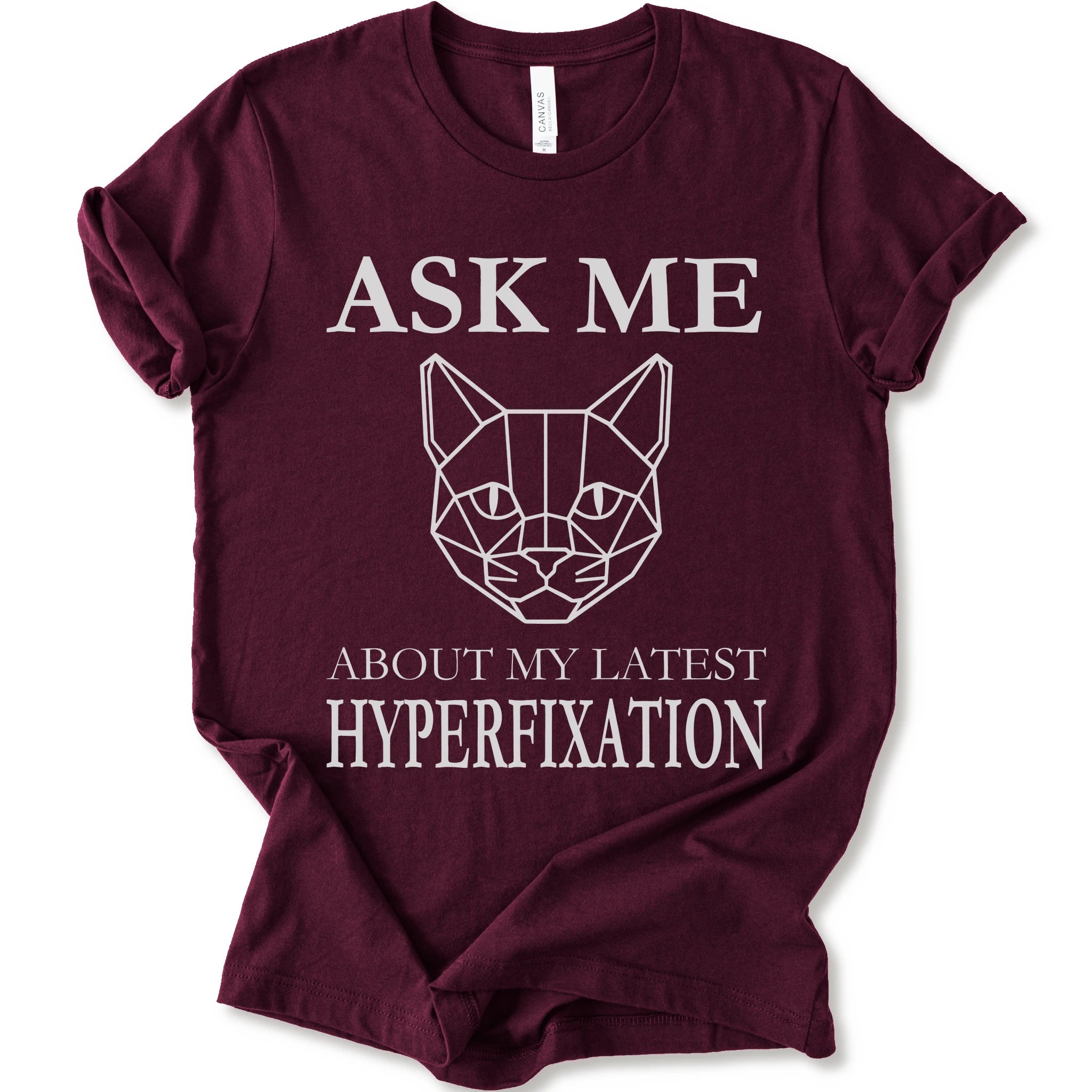 A maroon short-sleeve t-shirt with a white graphic print featuring a geometric cat face and the text 'ASK ME ABOUT MY LATEST HYPERFIXATION'.
