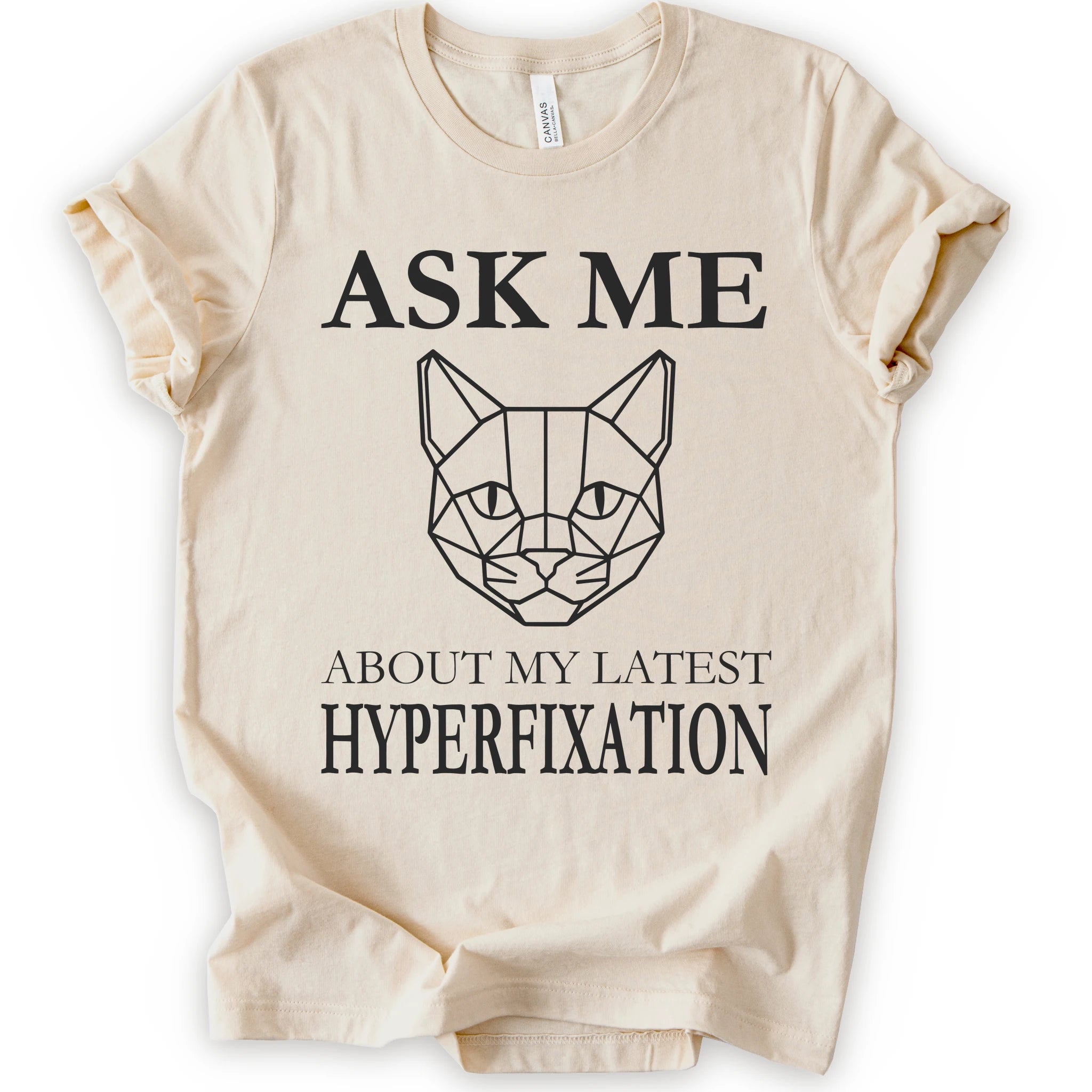 A natural short-sleeve t-shirt with a black graphic print featuring a geometric cat face and the text 'ASK ME ABOUT MY LATEST HYPERFIXATION'.
