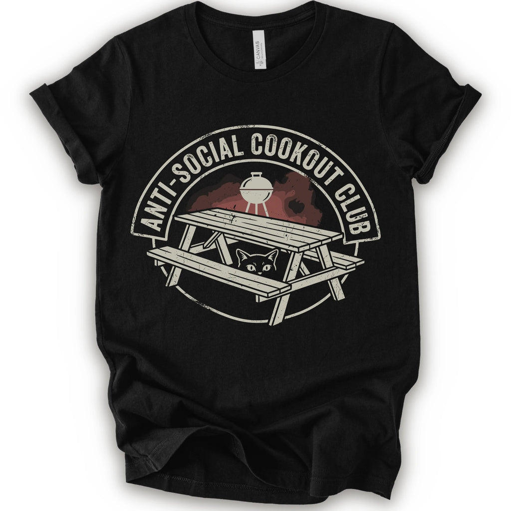A black short-sleeve crewneck t-shirt with a distressed graphic on the front. The graphic features a picnic table, a grill, and a cat peeking out from underneath, enclosed in a circle with the text 'ANTI-SOCIAL COOKOUT CLUB'.