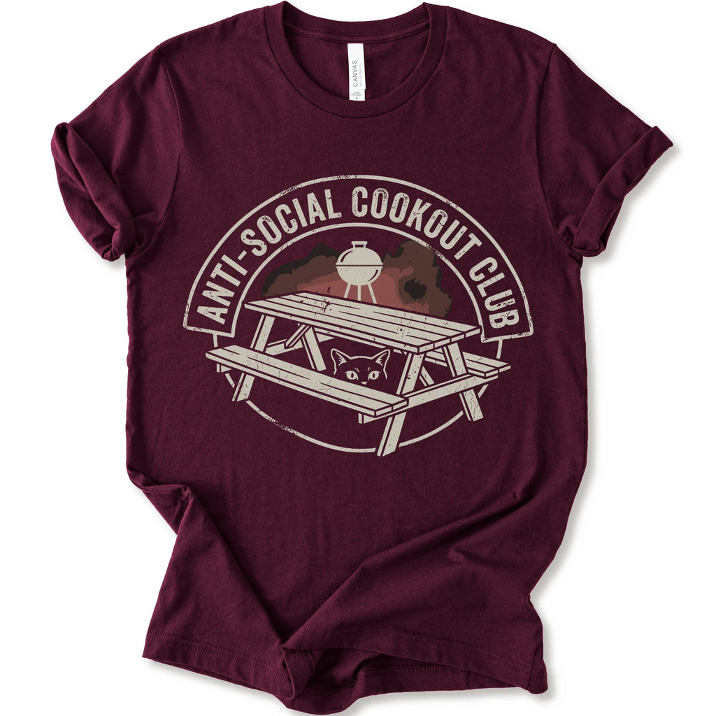 A maroon short-sleeve crewneck t-shirt with a distressed graphic on the front. The graphic features a picnic table, a grill, and a cat peeking out from underneath, enclosed in a circle with the text 'ANTI-SOCIAL COOKOUT CLUB'.
