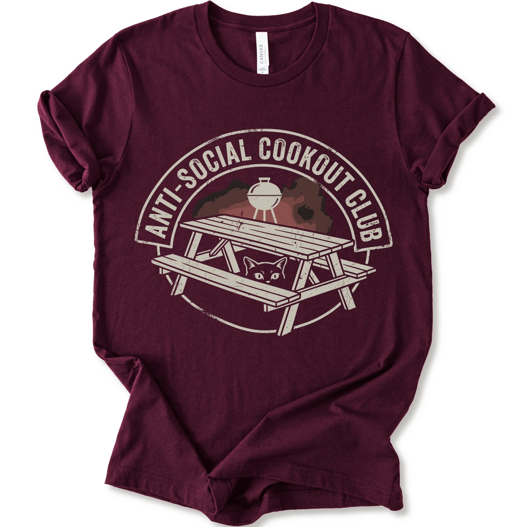 A maroon short-sleeve crewneck t-shirt with a distressed graphic on the front. The graphic features a picnic table, a grill, and a cat peeking out from underneath, enclosed in a circle with the text 'ANTI-SOCIAL COOKOUT CLUB'.