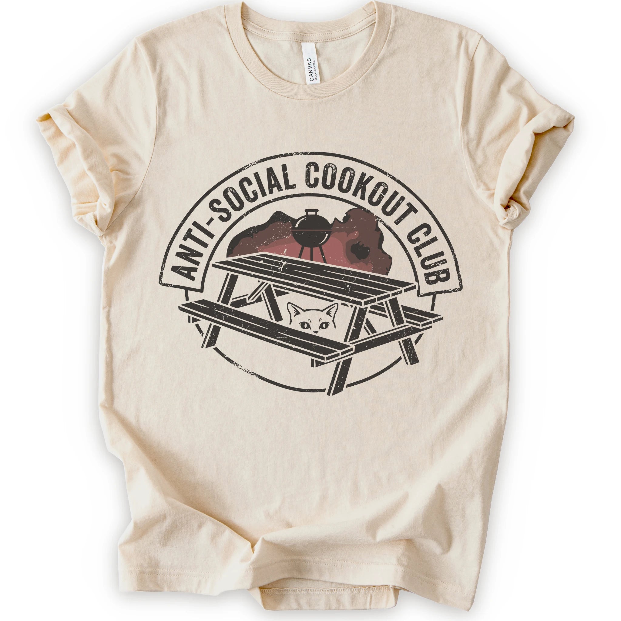 A natural short-sleeve crewneck t-shirt with a distressed graphic on the front. The graphic features a picnic table, a grill, and a cat peeking out from underneath, enclosed in a circle with the text 'ANTI-SOCIAL COOKOUT CLUB'.