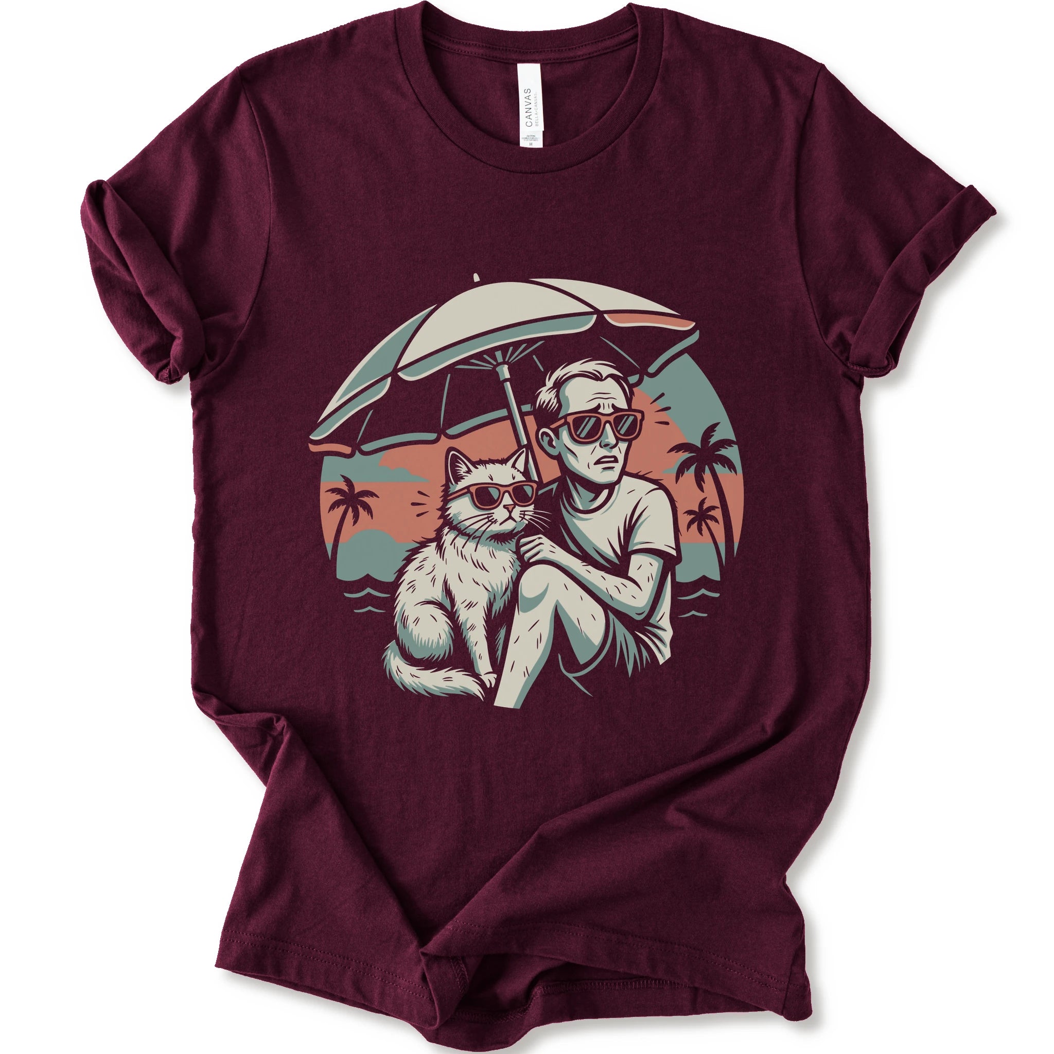 A maroon short-sleeve crewneck t-shirt with a vintage-style graphic print of a man and a cat sitting under a beach umbrella on a sunset beach.