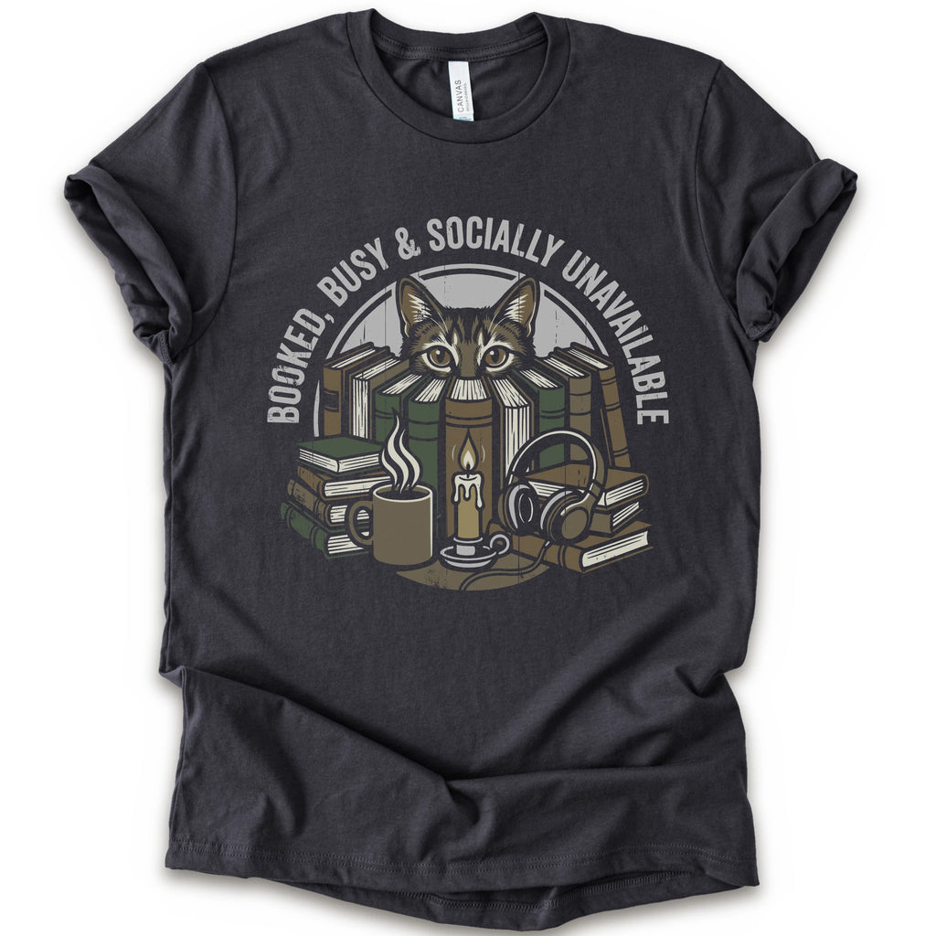 A dark grey short-sleeve crewneck t-shirt with a graphic print of a cat peeking out from stacked books, headphones, and a mug, with the text 'BOOKED BUSY & SOCIALLY UNAVAILABLE' arched above.