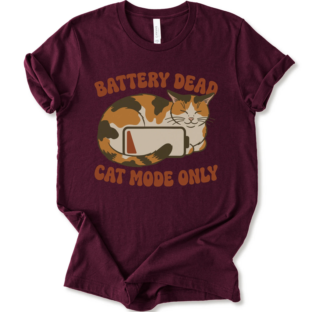 A maroon short-sleeve graphic t-shirt featuring a cartoon calico cat curled up around a battery icon, with the text 'BATTERY DEAD CAT MODE ONLY' in orange-brown lettering.