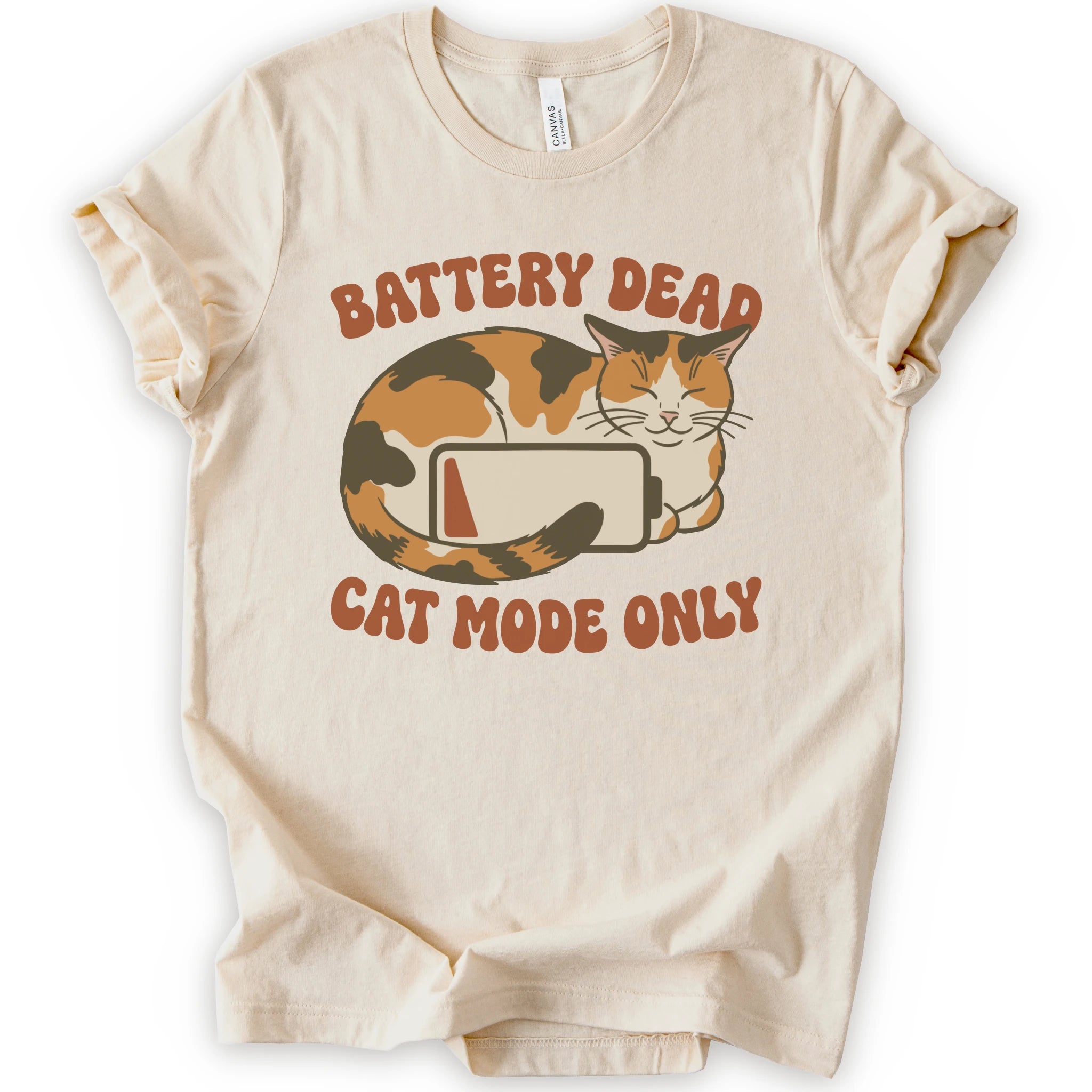 A natural short-sleeve graphic t-shirt featuring a cartoon calico cat curled up around a battery icon, with the text 'BATTERY DEAD CAT MODE ONLY' in orange-brown lettering.