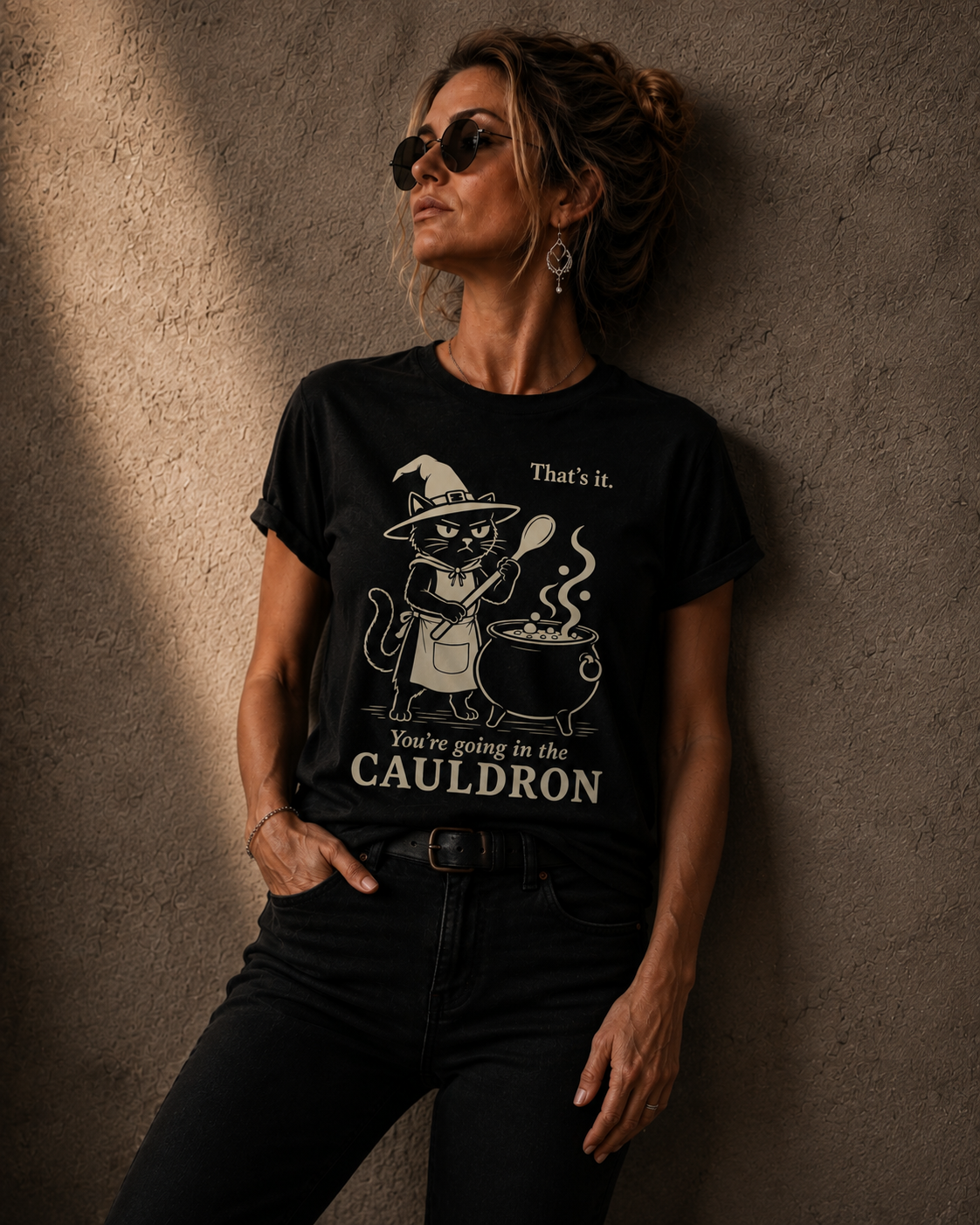 A woman wearing a black graphic t-shirt with a witch cat and cauldron design, leaning against a textured concrete wall.