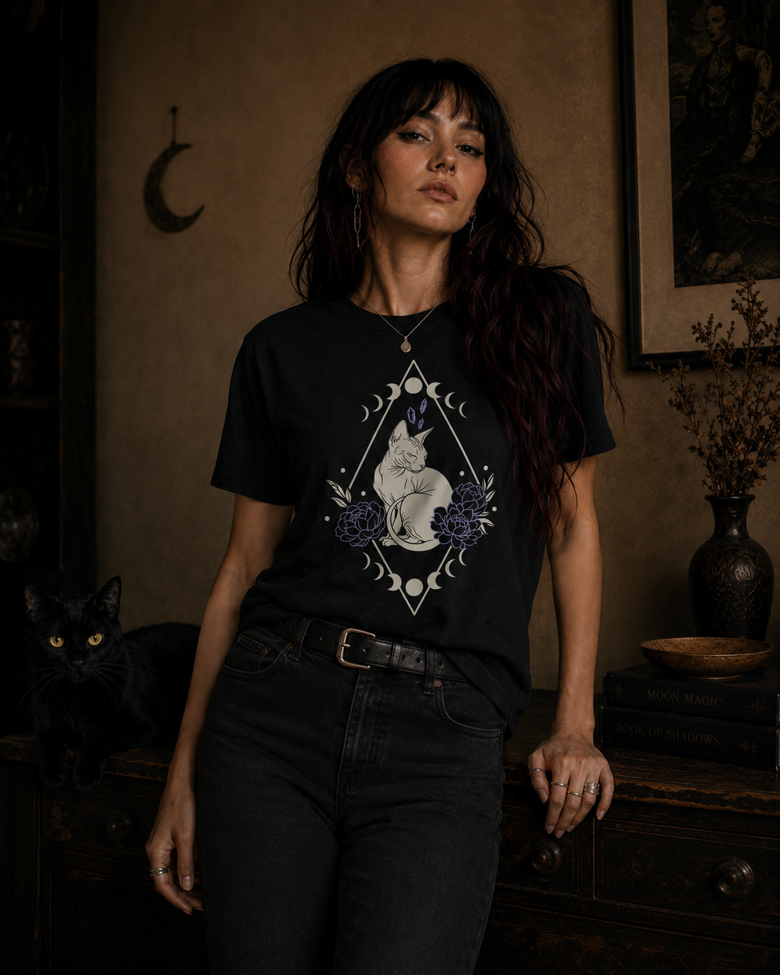 A woman wearing a black graphic t-shirt with a white sphynx cat and moon design, paired with dark jeans, standing in a dimly lit room with a black cat on a table beside her.