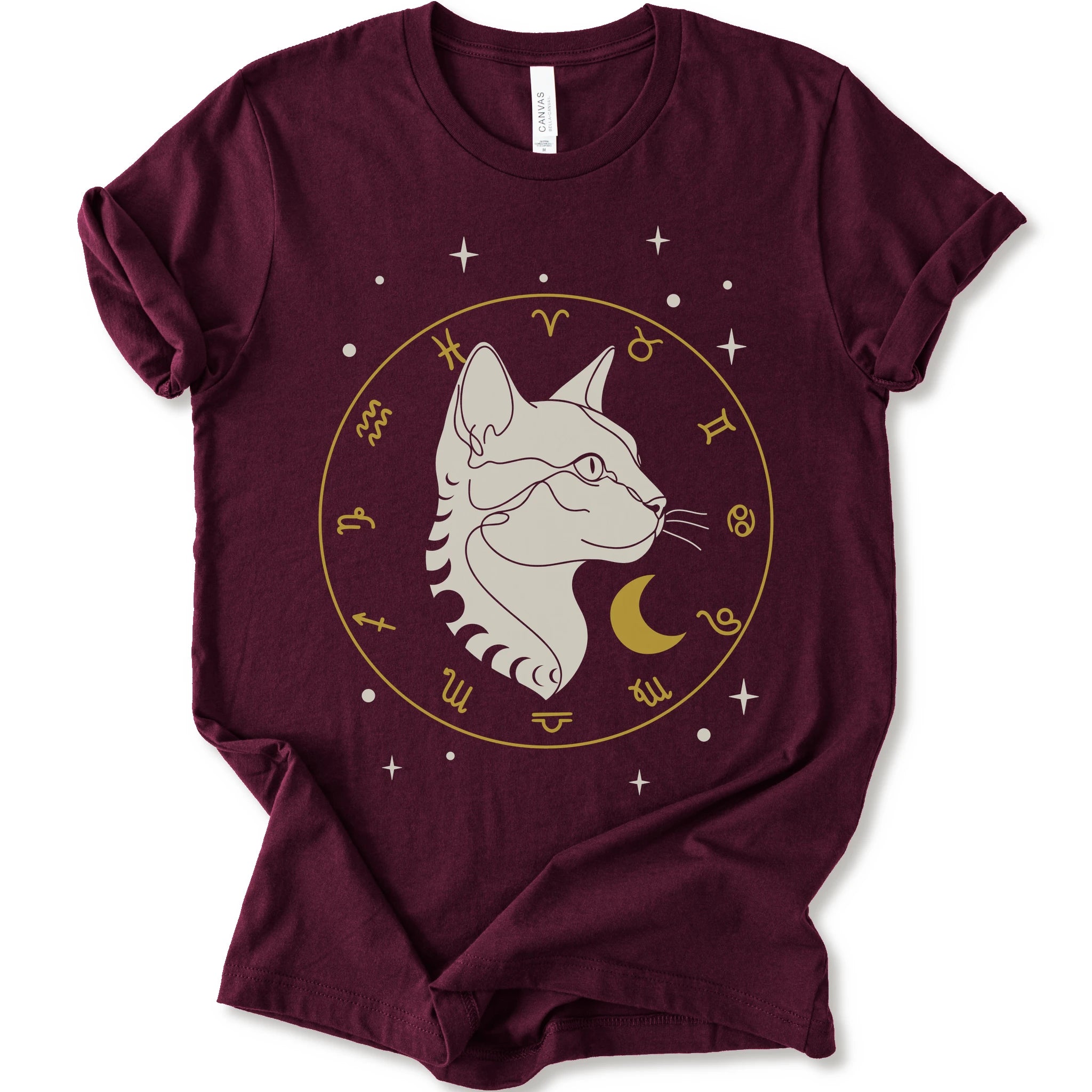 A maroon short-sleeve crewneck t-shirt featuring a graphic of a white cat profile inside a gold zodiac wheel with a crescent moon and stars.
