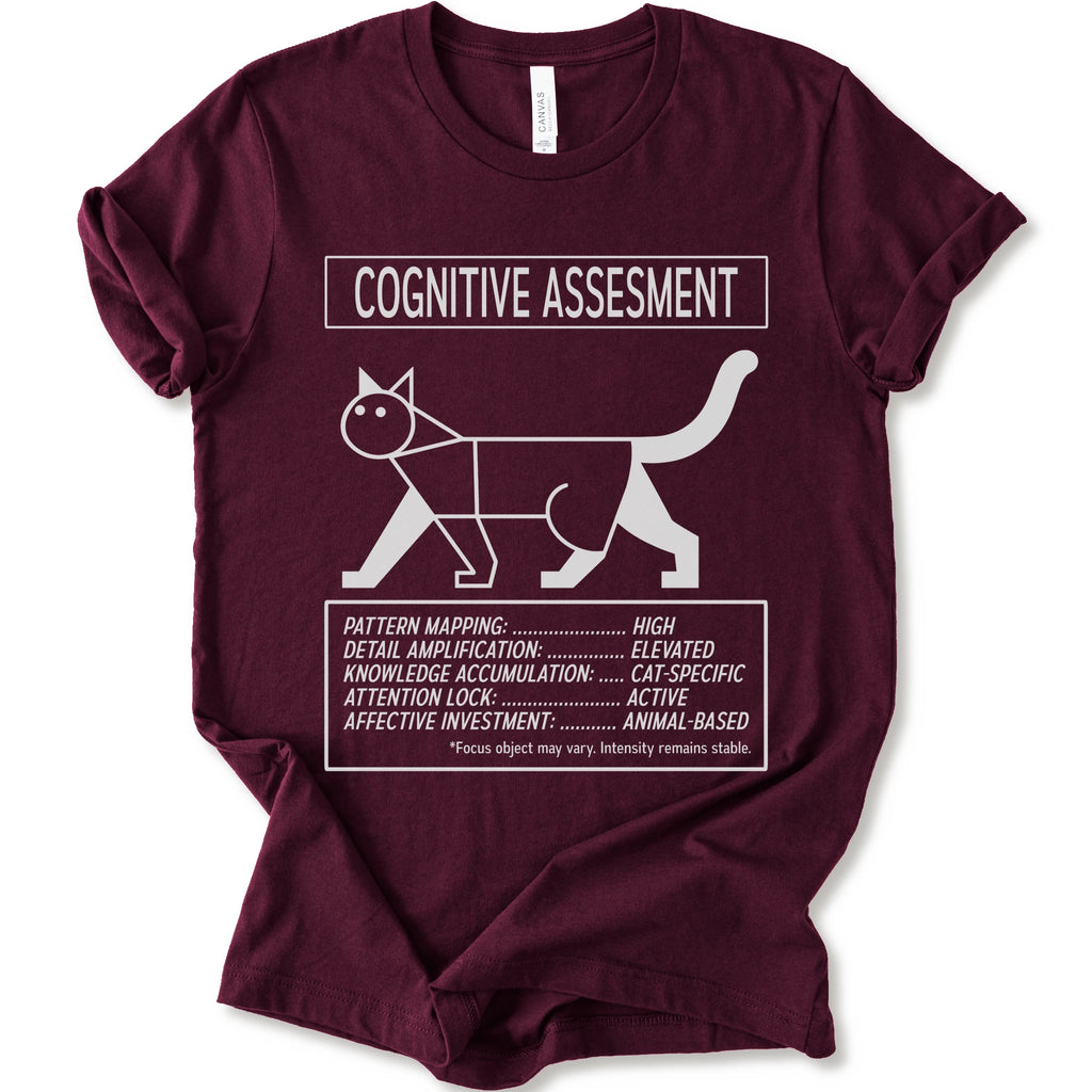 A maroon short-sleeve t-shirt with a white graphic print titled 'COGNITIVE ASSESSMENT' featuring a minimalist line drawing of a cat and a checklist-style design.