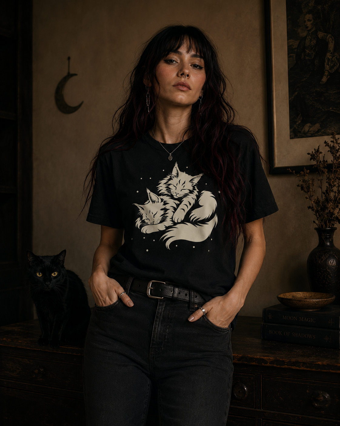A woman stands in a dimly lit room wearing a black graphic T-shirt with a white illustration of two maine coon cats, paired with dark jeans. A black cat sits on a wooden cabinet to her left, and framed wall art and dried flowers are visible in the background.