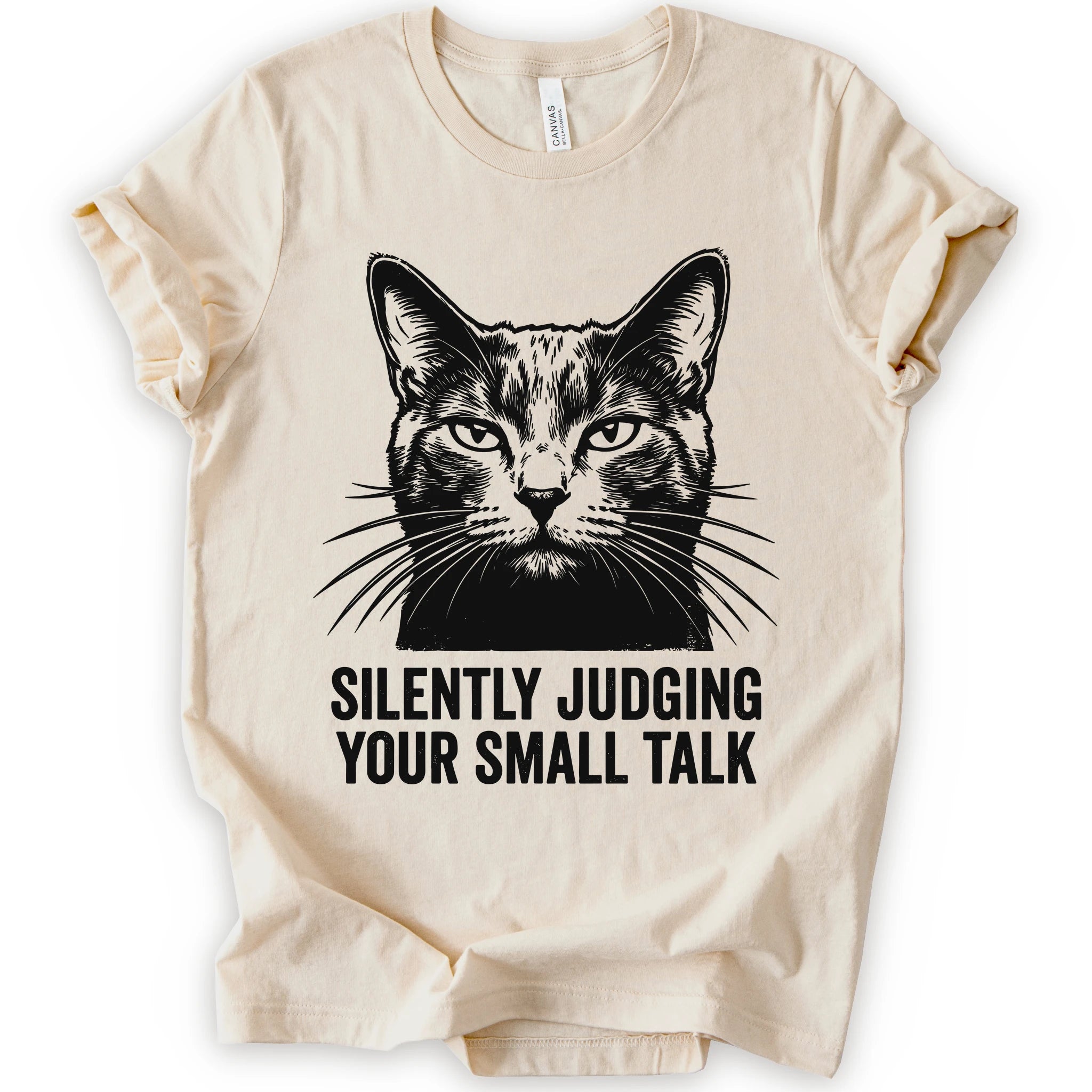 An natural short-sleeve t-shirt with a black graphic of a cat's face and the text 'SILENTLY JUDGING YOUR SMALL TALK' printed on the front.