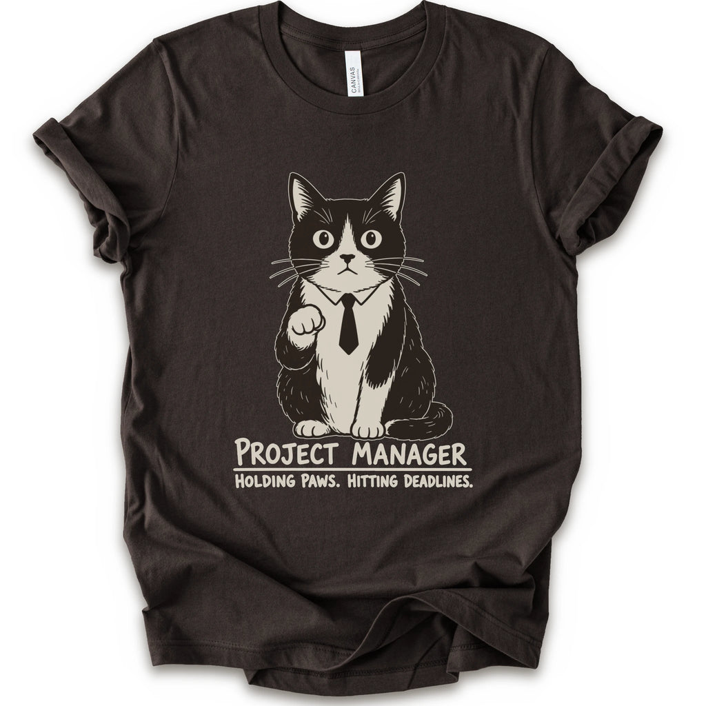 A brown short-sleeve crewneck t-shirt featuring a graphic of a black and white cat wearing a tie with the text 'PROJECT MANAGER HOLDING PAWS. HITTING DEADLINES.
