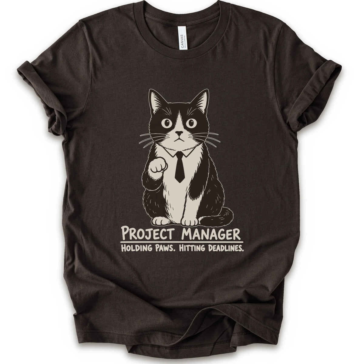 A brown short-sleeve crewneck t-shirt featuring a graphic of a black and white cat wearing a tie with the text 'PROJECT MANAGER HOLDING PAWS. HITTING DEADLINES.