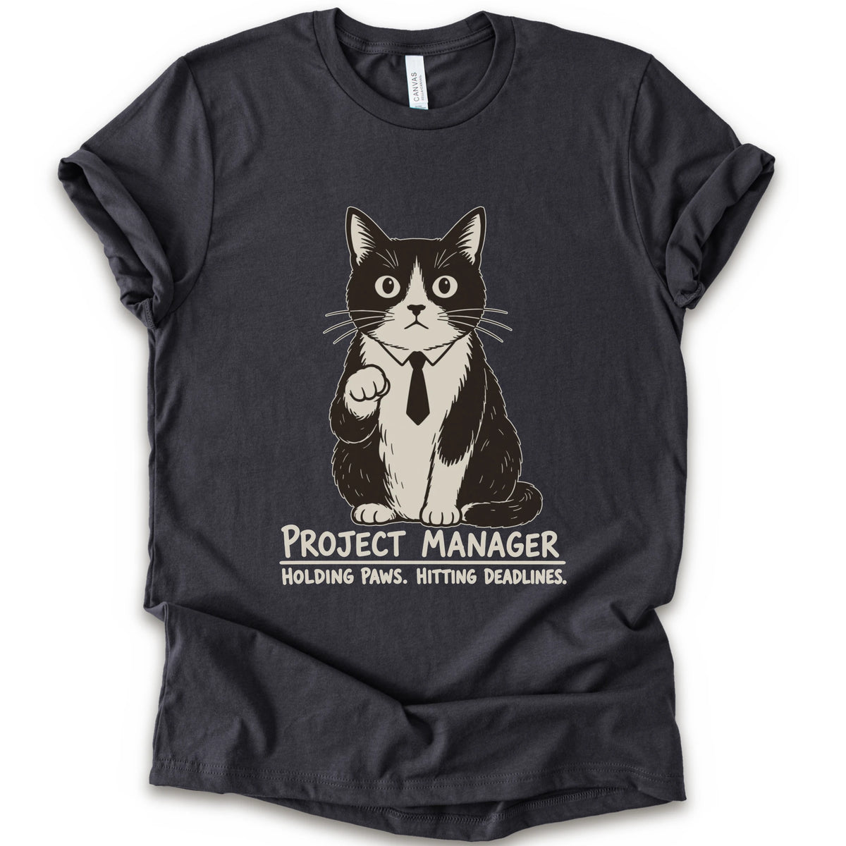 A dark grey short-sleeve crewneck t-shirt featuring a graphic of a black and white cat wearing a tie with the text 'PROJECT MANAGER HOLDING PAWS. HITTING DEADLINES.