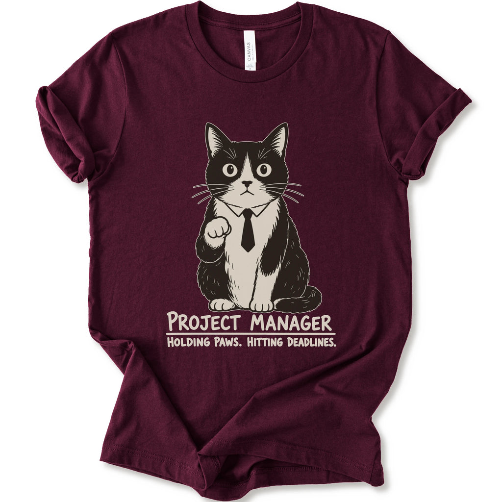A maroon short-sleeve crewneck t-shirt featuring a graphic of a black and white cat wearing a tie with the text 'PROJECT MANAGER HOLDING PAWS. HITTING DEADLINES.