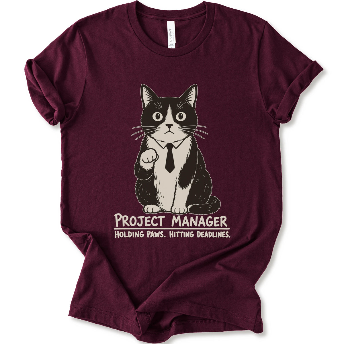 A maroon short-sleeve crewneck t-shirt featuring a graphic of a black and white cat wearing a tie with the text 'PROJECT MANAGER HOLDING PAWS. HITTING DEADLINES.