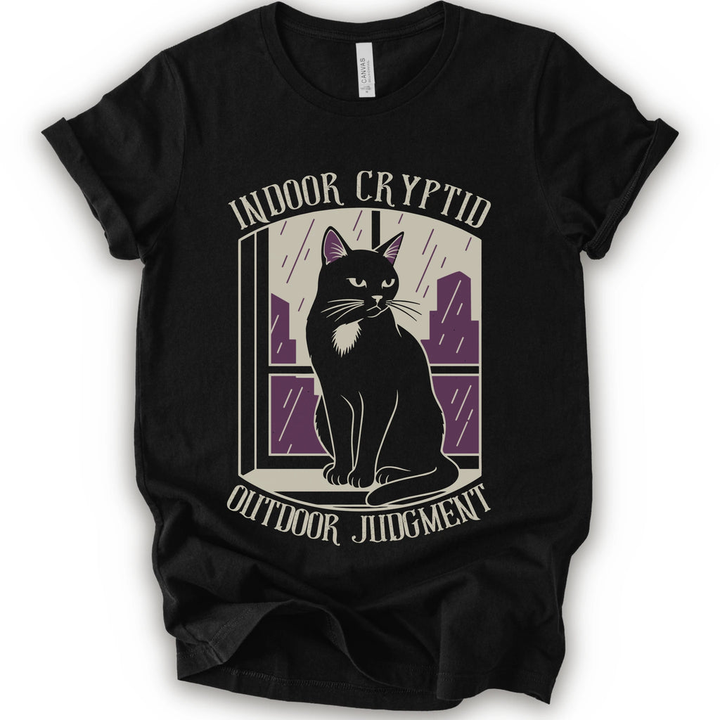 A black short-sleeve graphic t-shirt featuring a stylized illustration of a black cat sitting in front of a window with purple rain streaks, with the text 'INDOOR CRYPTID' and 'OUTDOOR JUDGMENT' printed on the front.