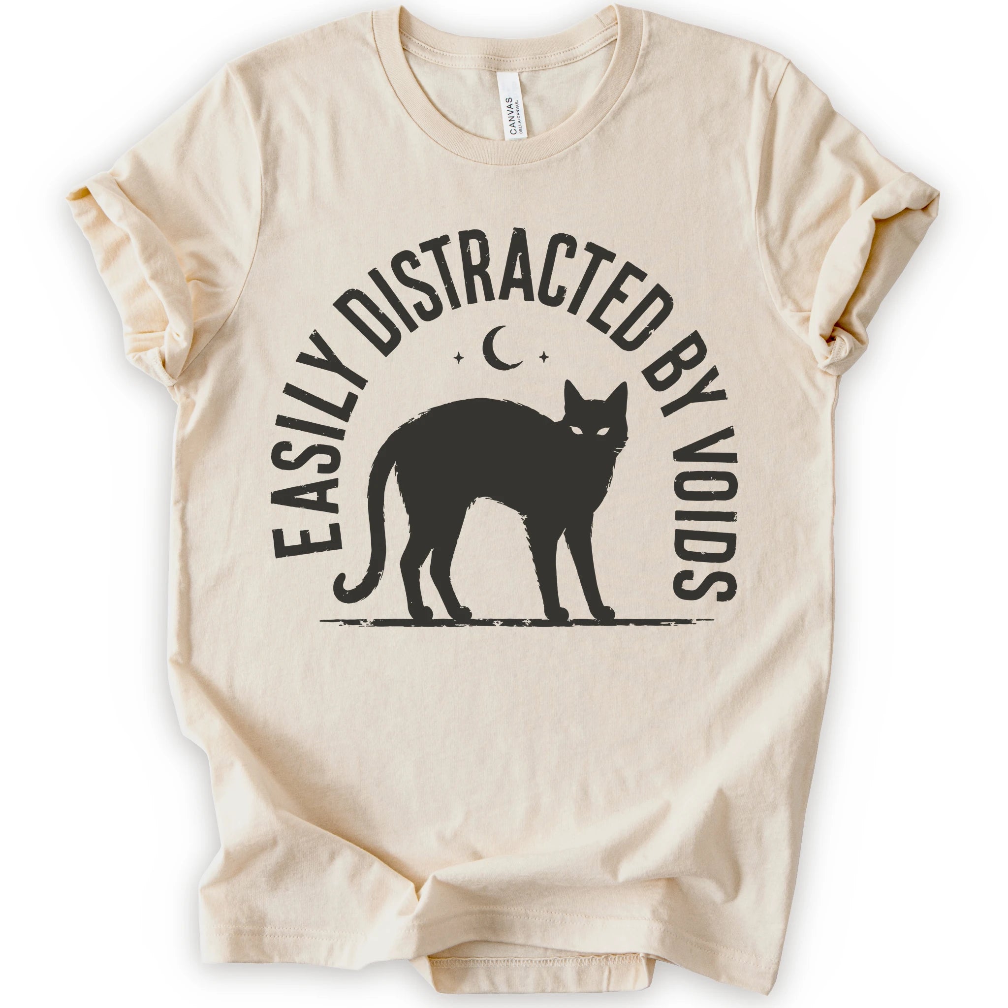 A natural short-sleeve t-shirt with a black graphic print of a cat silhouette, a crescent moon, and the text 'EASILY DISTRACTED BY VOIDS' on the front