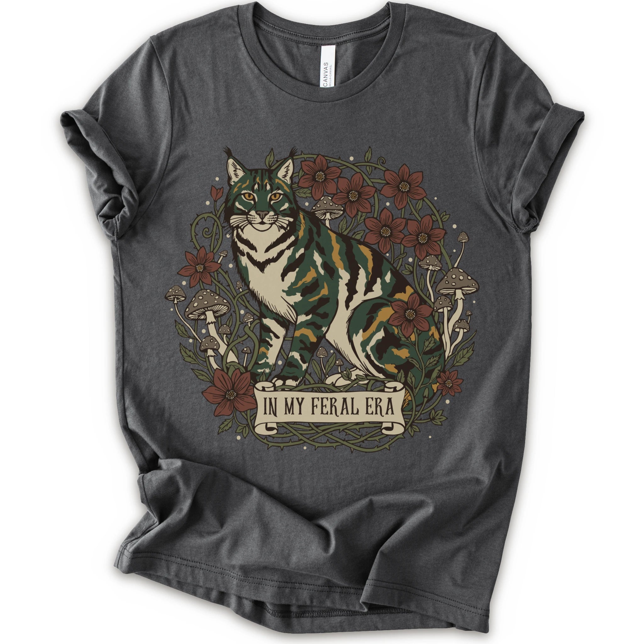 An asphalt short-sleeve crewneck t-shirt featuring a detailed graphic of a green, yellow, brown and white cat surrounded by red flowers, mushrooms, and vines, with a banner reading 'IN MY FERAL ERA'.