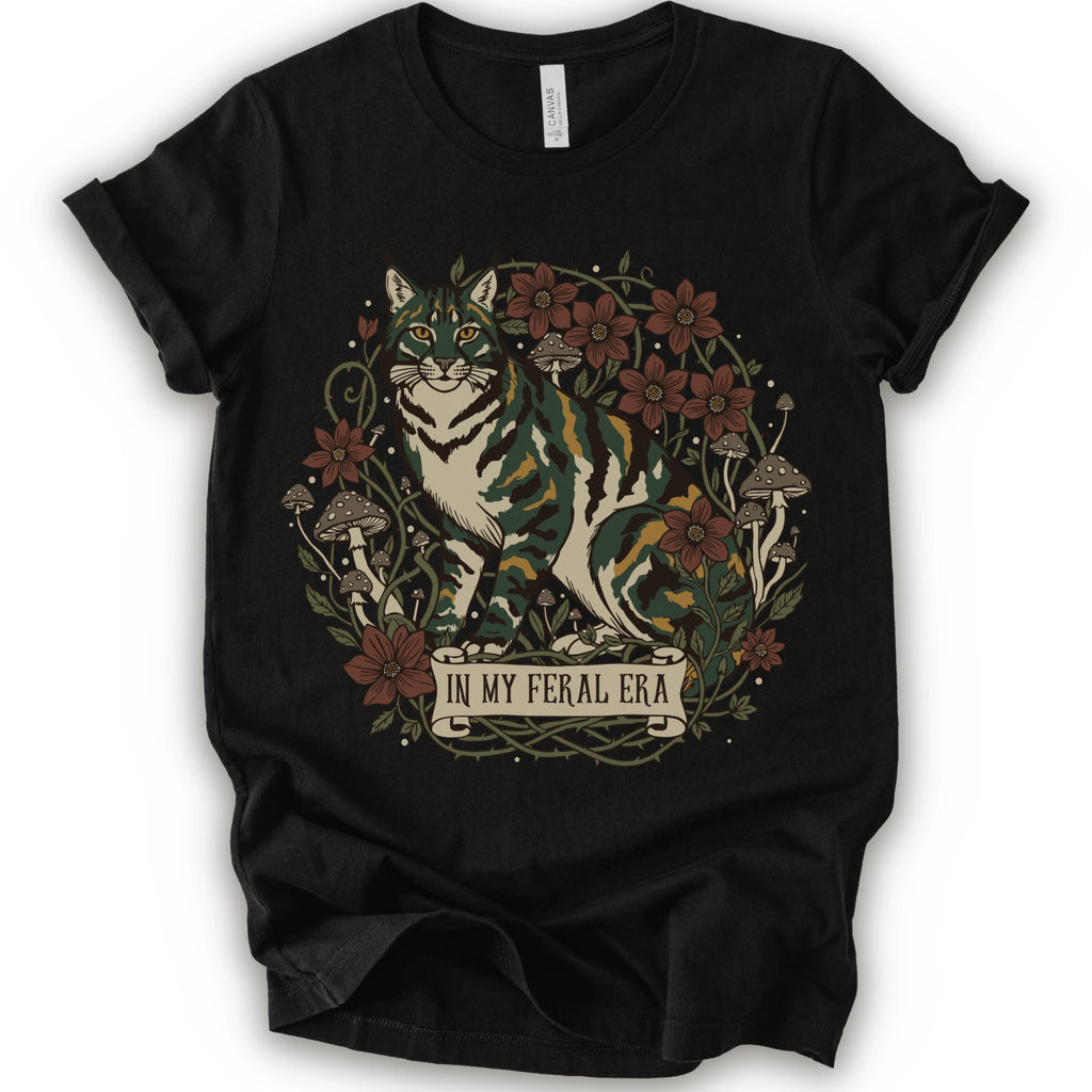 A black short-sleeve crewneck t-shirt featuring a detailed graphic of a green, yellow, brown and white cat surrounded by red flowers, mushrooms, and vines, with a banner reading 'IN MY FERAL ERA'.