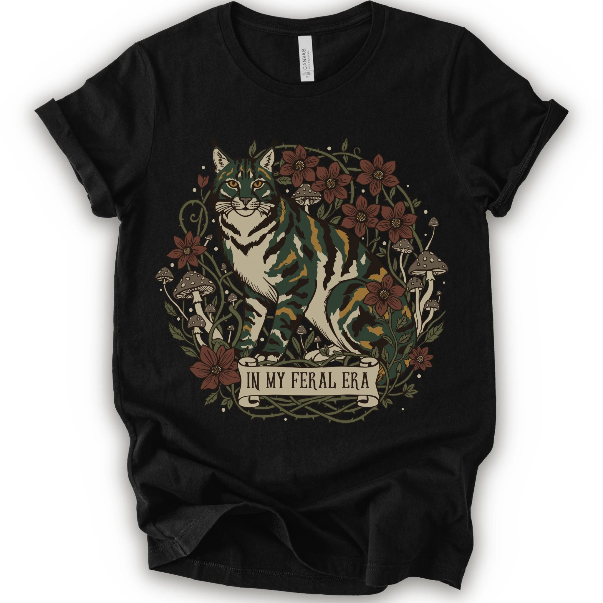 A black short-sleeve crewneck t-shirt featuring a detailed graphic of a green, yellow, brown and white cat surrounded by red flowers, mushrooms, and vines, with a banner reading 'IN MY FERAL ERA'.