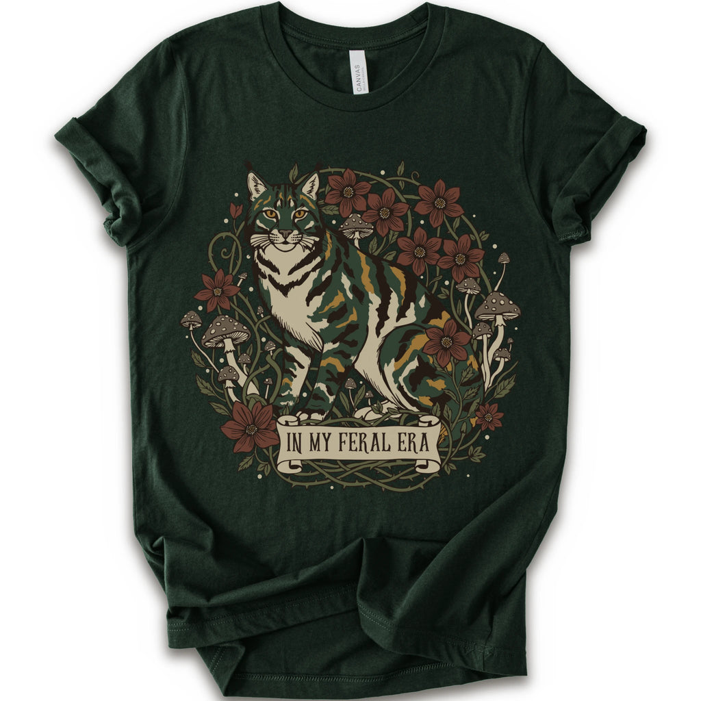 A forest short-sleeve crewneck t-shirt featuring a detailed graphic of a green, yellow, brown and white cat surrounded by red flowers, mushrooms, and vines, with a banner reading 'IN MY FERAL ERA'.