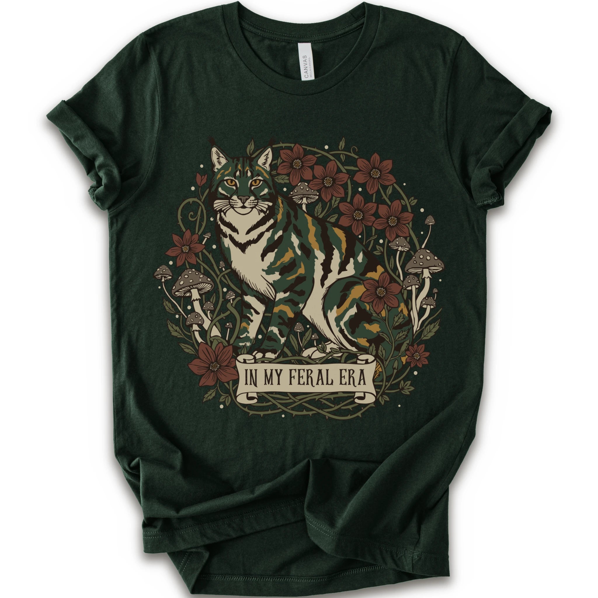 A forest short-sleeve crewneck t-shirt featuring a detailed graphic of a green, yellow, brown and white cat surrounded by red flowers, mushrooms, and vines, with a banner reading 'IN MY FERAL ERA'.
