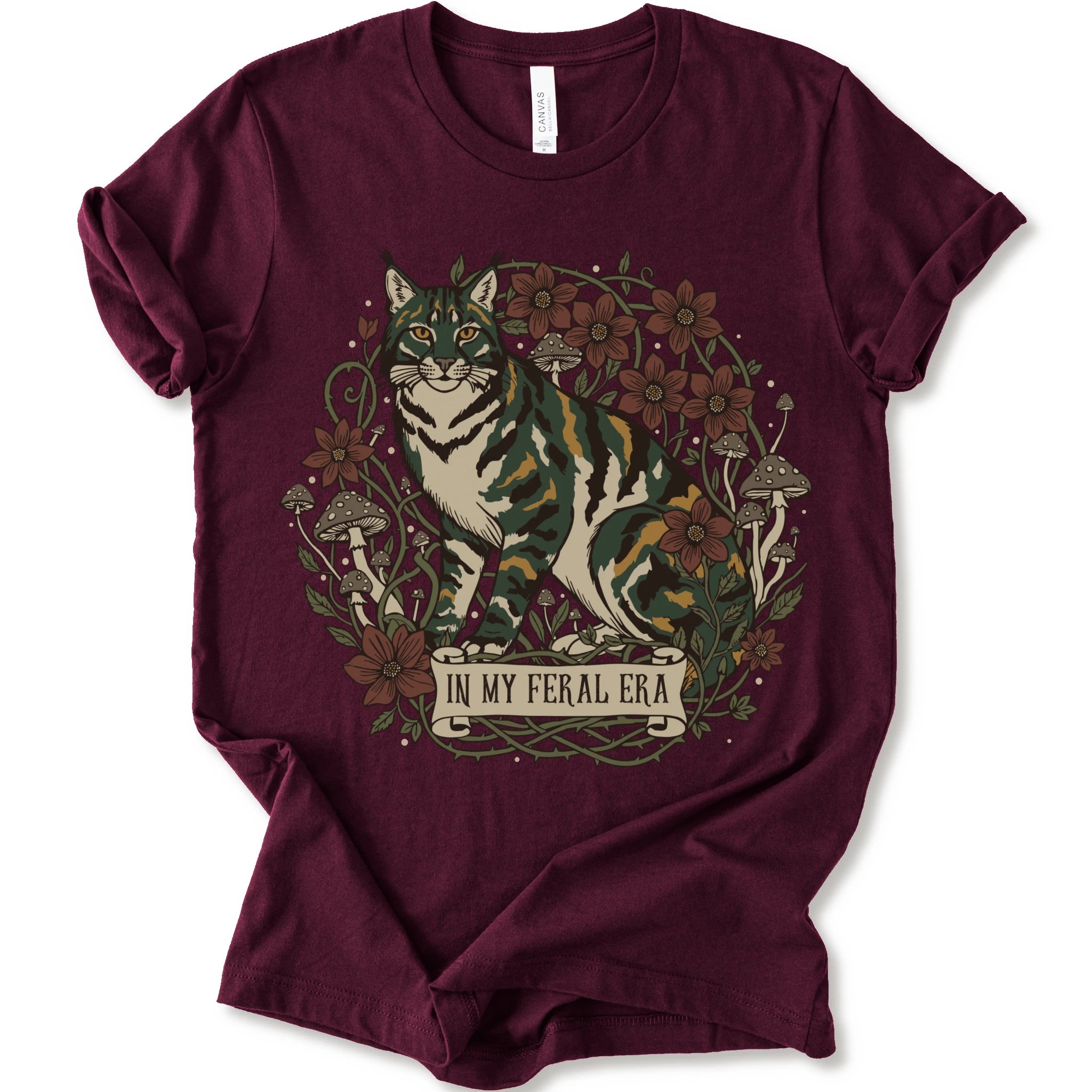 A maroon short-sleeve crewneck t-shirt featuring a detailed graphic of a green, yellow, brown and white cat surrounded by red flowers, mushrooms, and vines, with a banner reading 'IN MY FERAL ERA'.