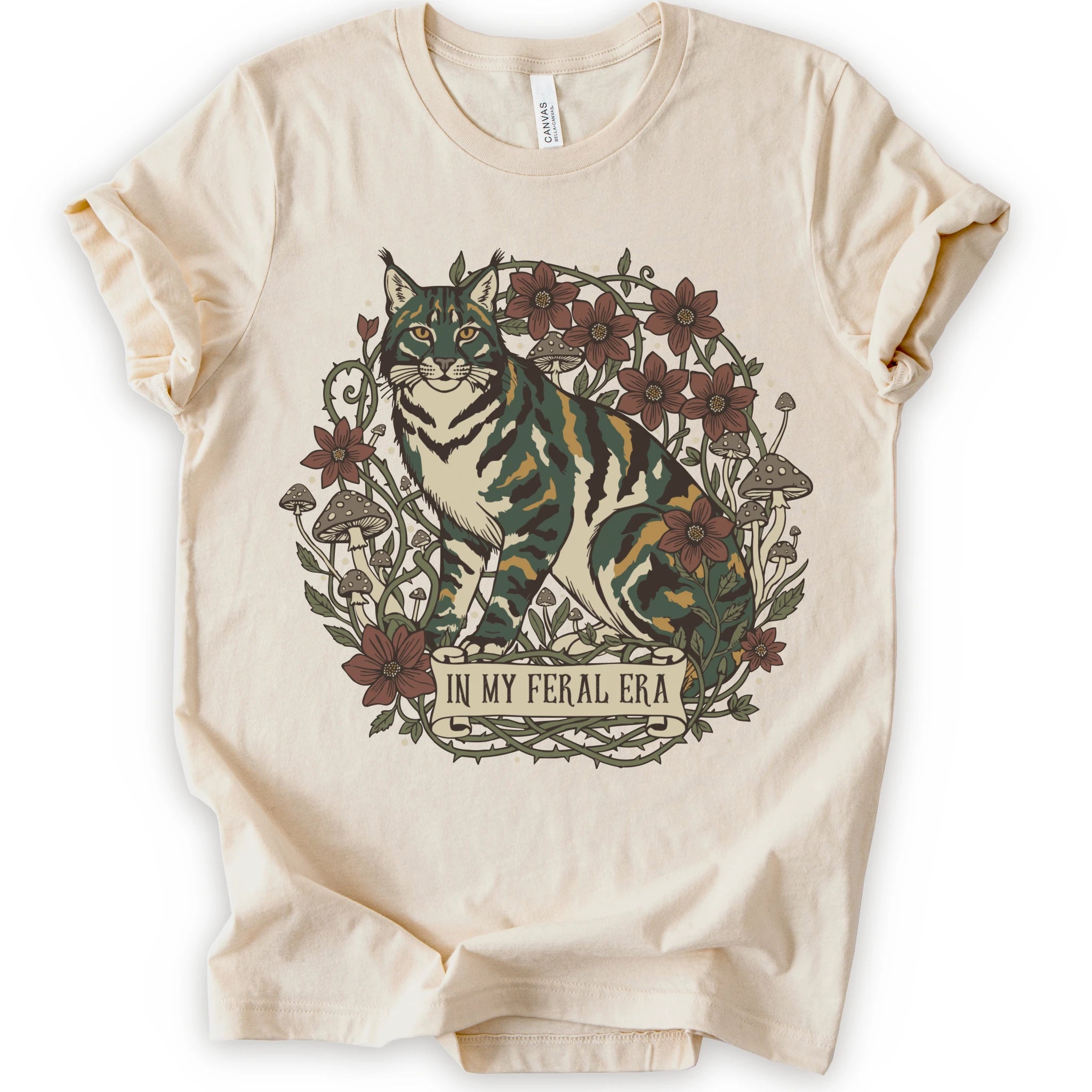 A natural short-sleeve crewneck t-shirt featuring a detailed graphic of a green, yellow, brown and white cat surrounded by red flowers, mushrooms, and vines, with a banner reading 'IN MY FERAL ERA'.