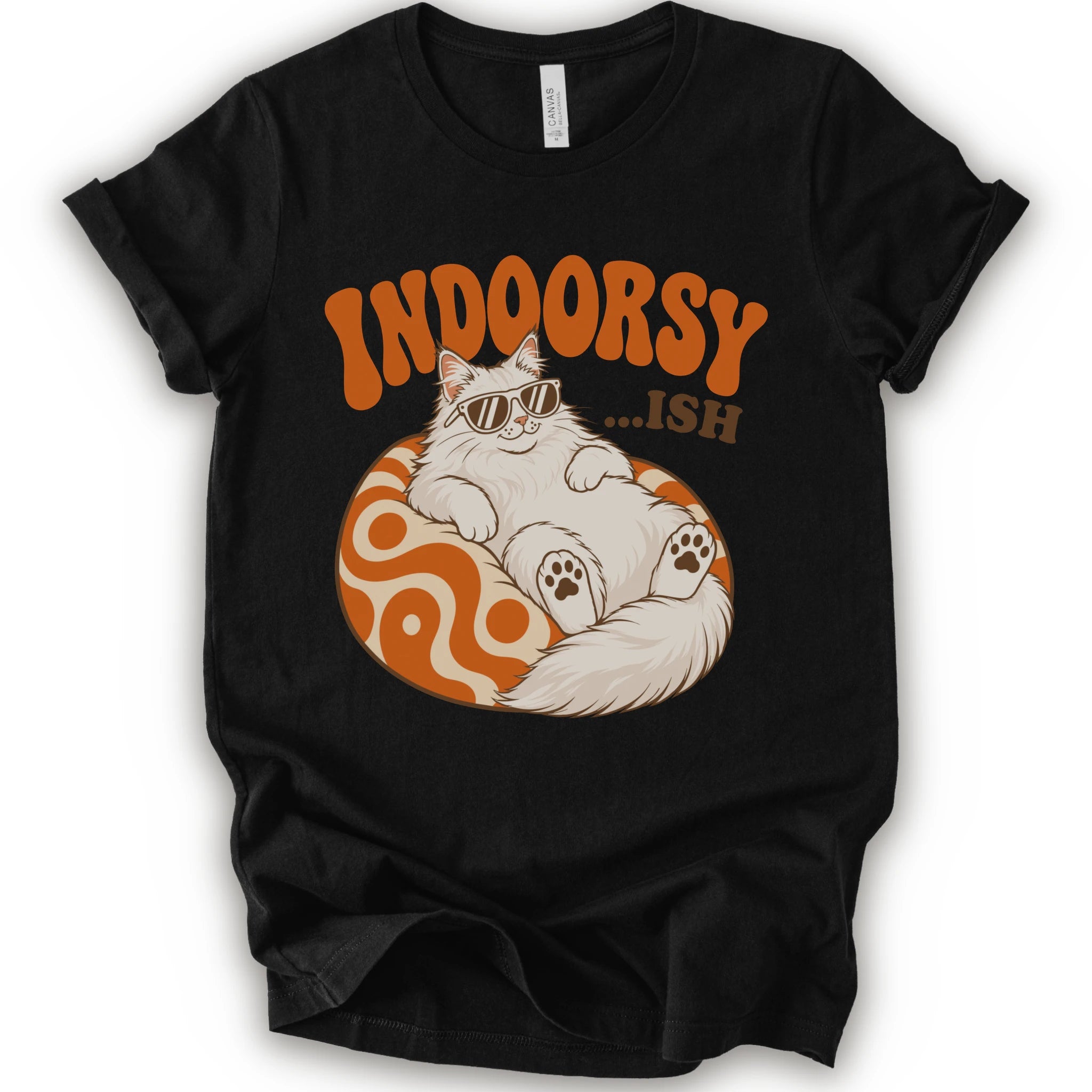 A black short-sleeve crewneck t-shirt with a graphic print of a fluffy white Maine Coon cat wearing sunglasses, lounging on an orange and white swirl-patterned sphere. Above the cat, the text reads 'INDOORSY...ish".