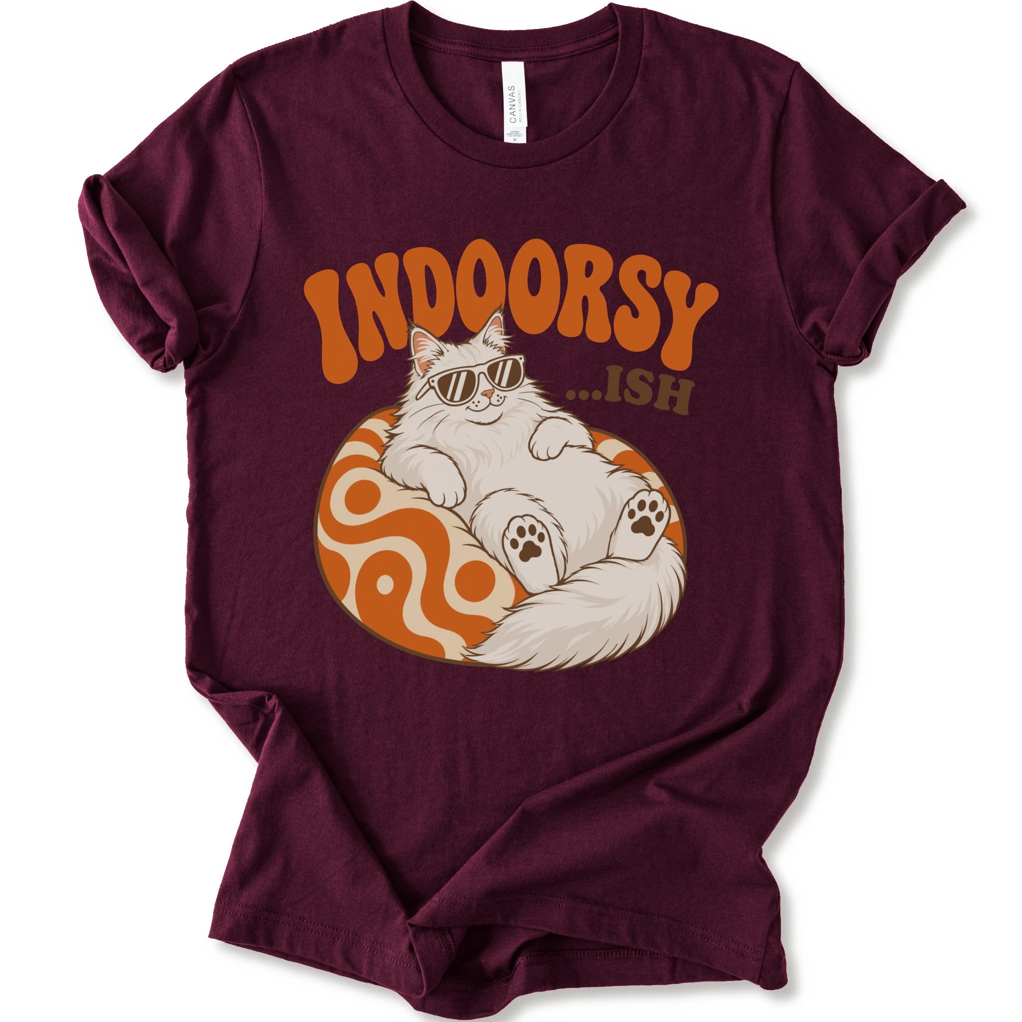 A maroon short-sleeve crewneck t-shirt with a graphic print of a fluffy white Maine Coon cat wearing sunglasses, lounging on an orange and white swirl-patterned sphere. Above the cat, the text reads 'INDOORSY...ish".