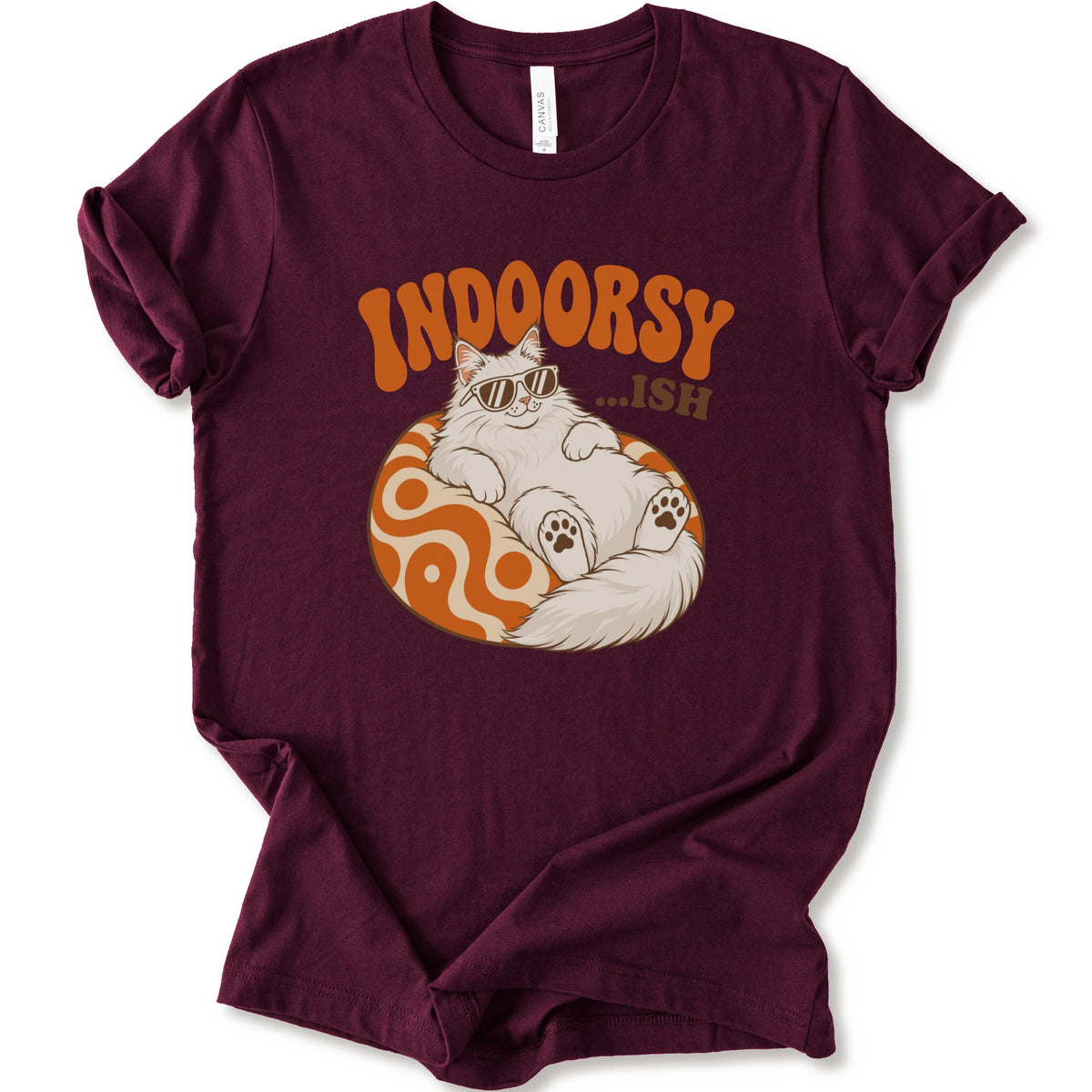 A maroon short-sleeve t-shirt with a graphic of a white cat wearing sunglasses on an orange patterned ball, with the text 'INDOORSY... .ish'.