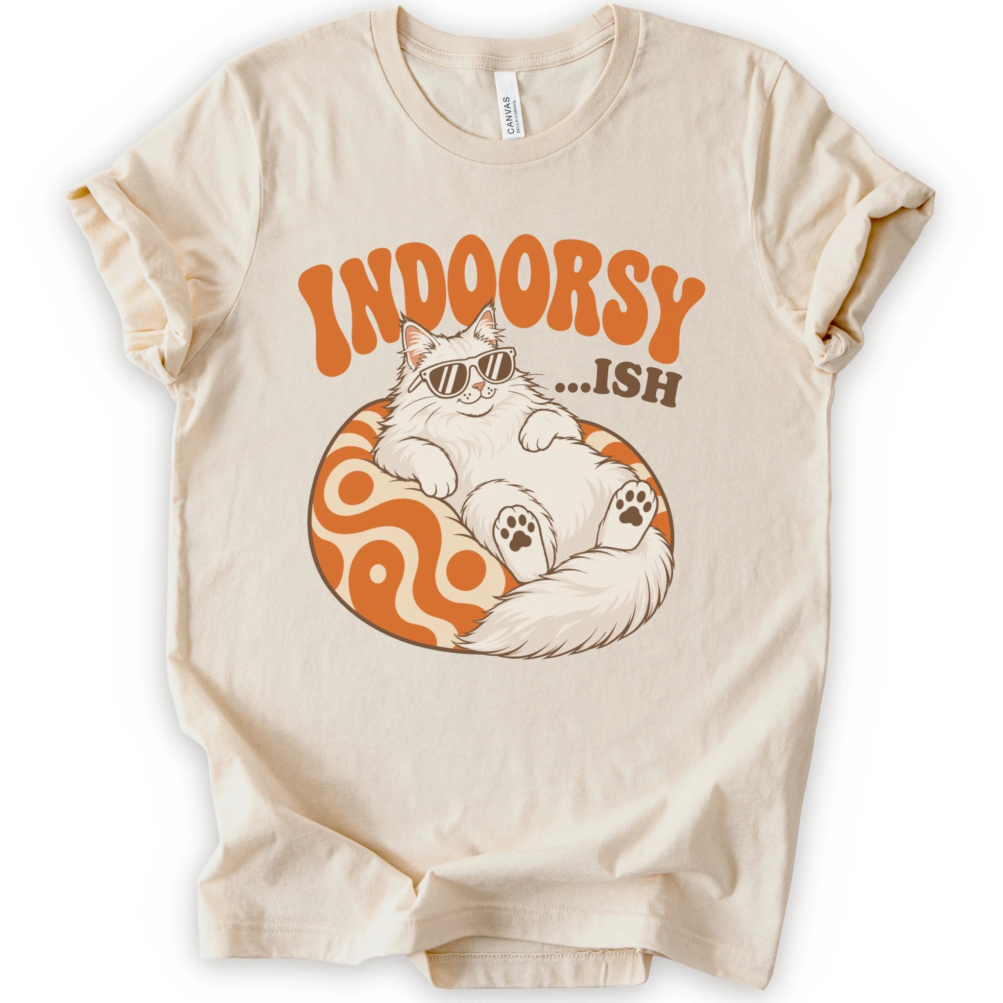 A natural short-sleeve crewneck t-shirt with a graphic print of a fluffy white Maine Coon cat wearing sunglasses, lounging on an orange and white swirl-patterned sphere. Above the cat, the text reads 'INDOORSY...ish".