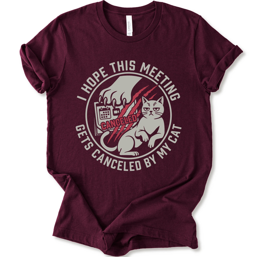 A maroon short-sleeve crewneck t-shirt with a circular graphic on the front. The graphic features a white cat, a hand scratching, and the text 'I HOPE THIS MEETING GETS CANCELLED BY MY CAT' in white, with 'CANCELLED' written in red script.
