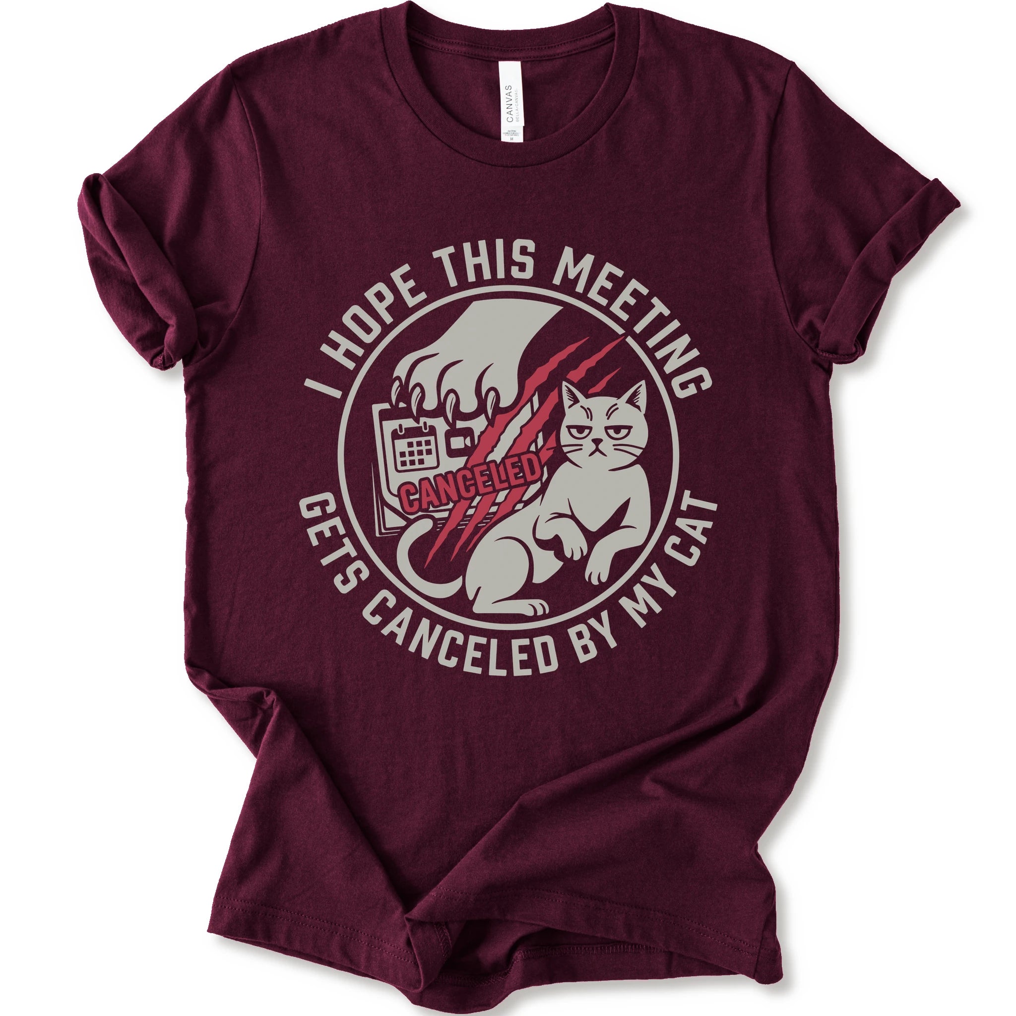 A maroon short-sleeve crewneck t-shirt with a circular graphic on the front. The graphic features a white cat, a hand scratching, and the text 'I HOPE THIS MEETING GETS CANCELLED BY MY CAT' in white, with 'CANCELLED' written in red script.