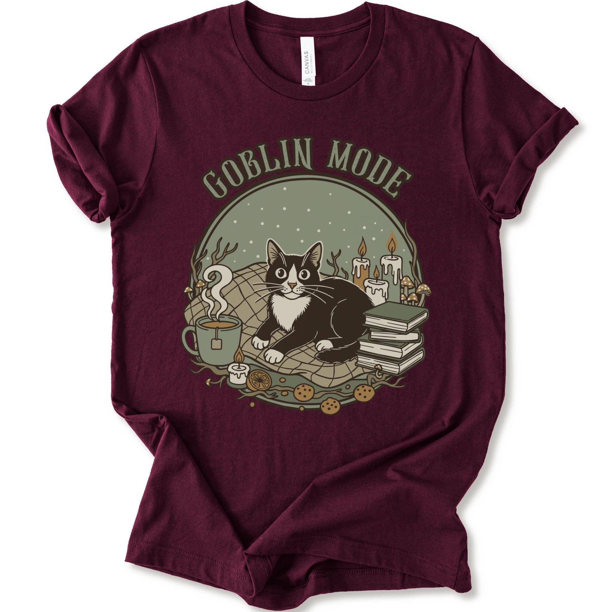 A maroon short-sleeve crewneck t-shirt with a cozy 'Goblin Mode' graphic print featuring a black and white cat, candles, books, and a mug.