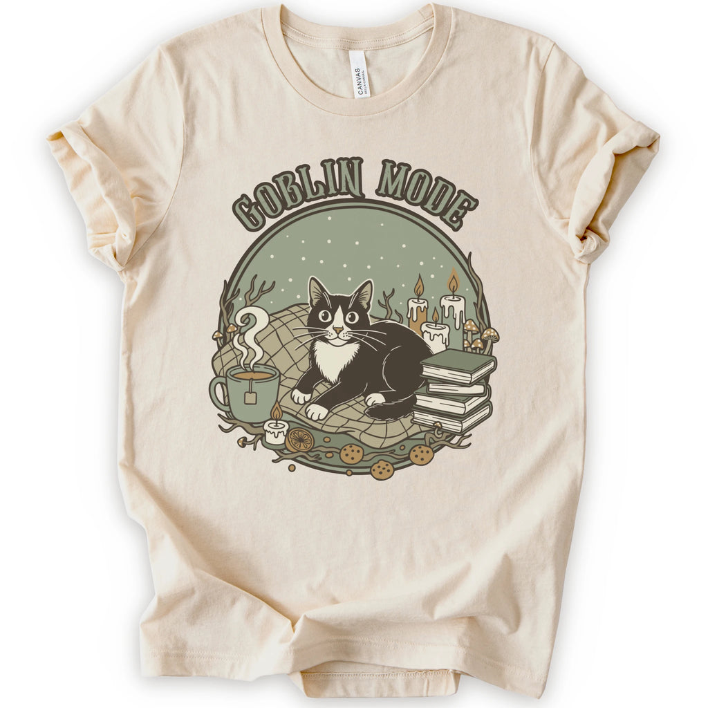 A natural short-sleeve crewneck t-shirt with a cozy 'Goblin Mode' graphic print featuring a black and white cat, candles, books, and a mug.