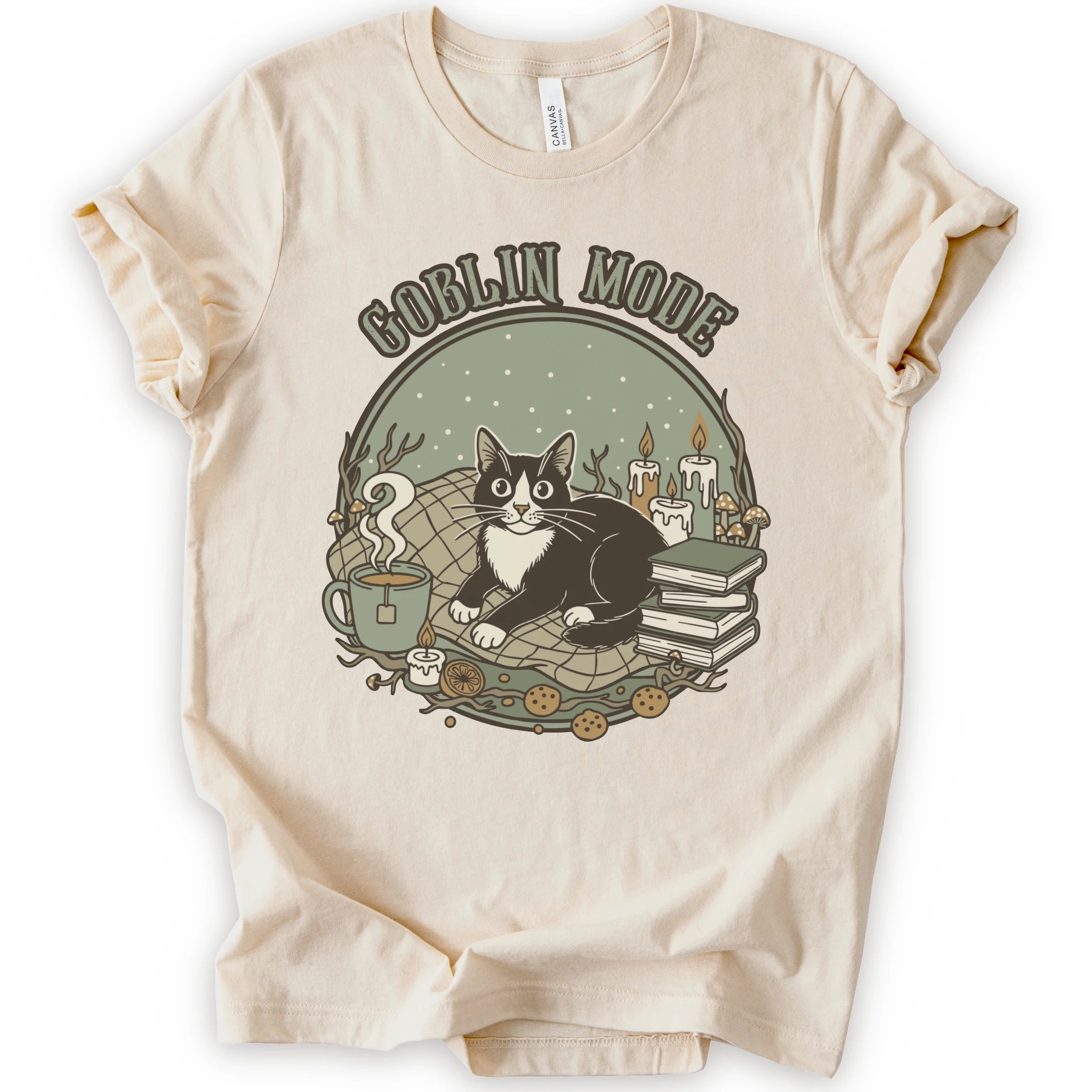 A natural short-sleeve crewneck t-shirt with a cozy 'Goblin Mode' graphic print featuring a black and white cat, candles, books, and a mug.