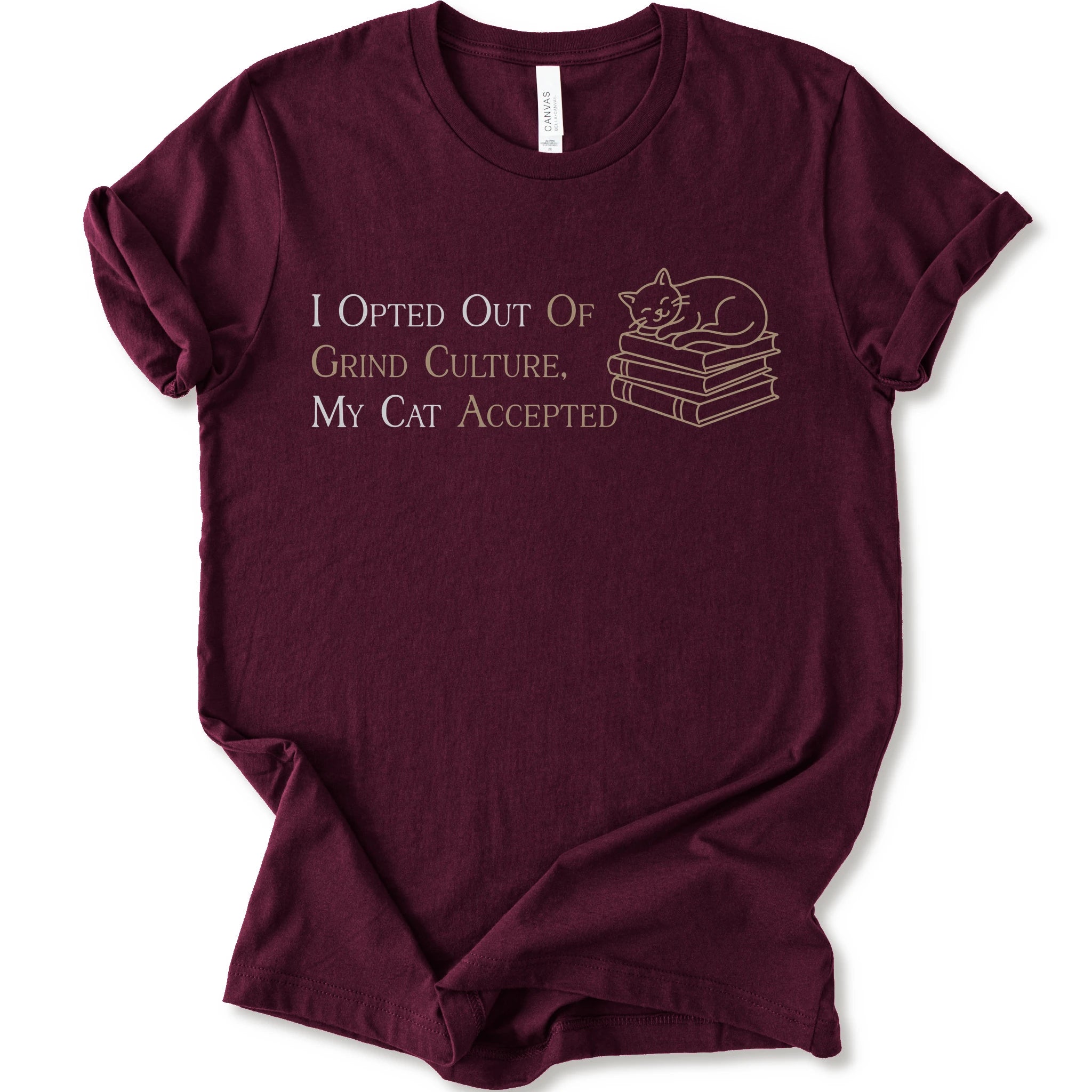 A maroon short-sleeve crew neck T-shirt with "I Opted Out Of Grind Culture, My Cat Accepted" lettering beside a sleeping cat on stacked books
