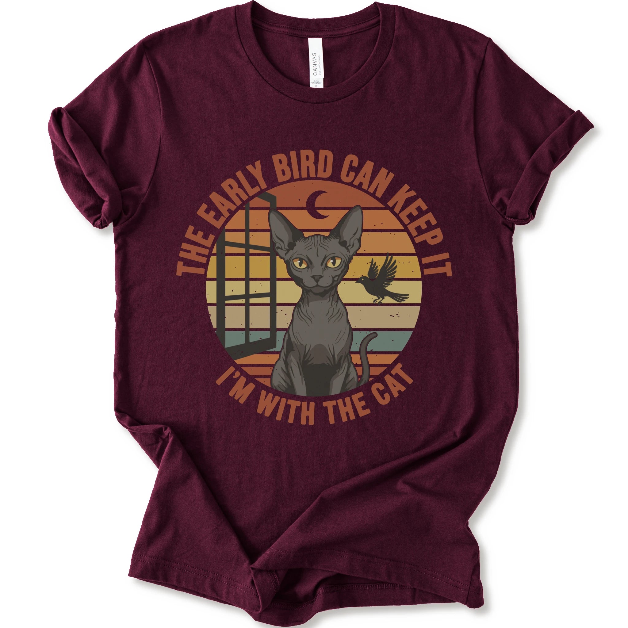 A maroon short-sleeve crewneck t-shirt with a graphic print of a hairless cat in front of a retro sunset circle, with the text 'THE EARLY BIRD CAN KEEP IT I'M WITH THE CAT'.