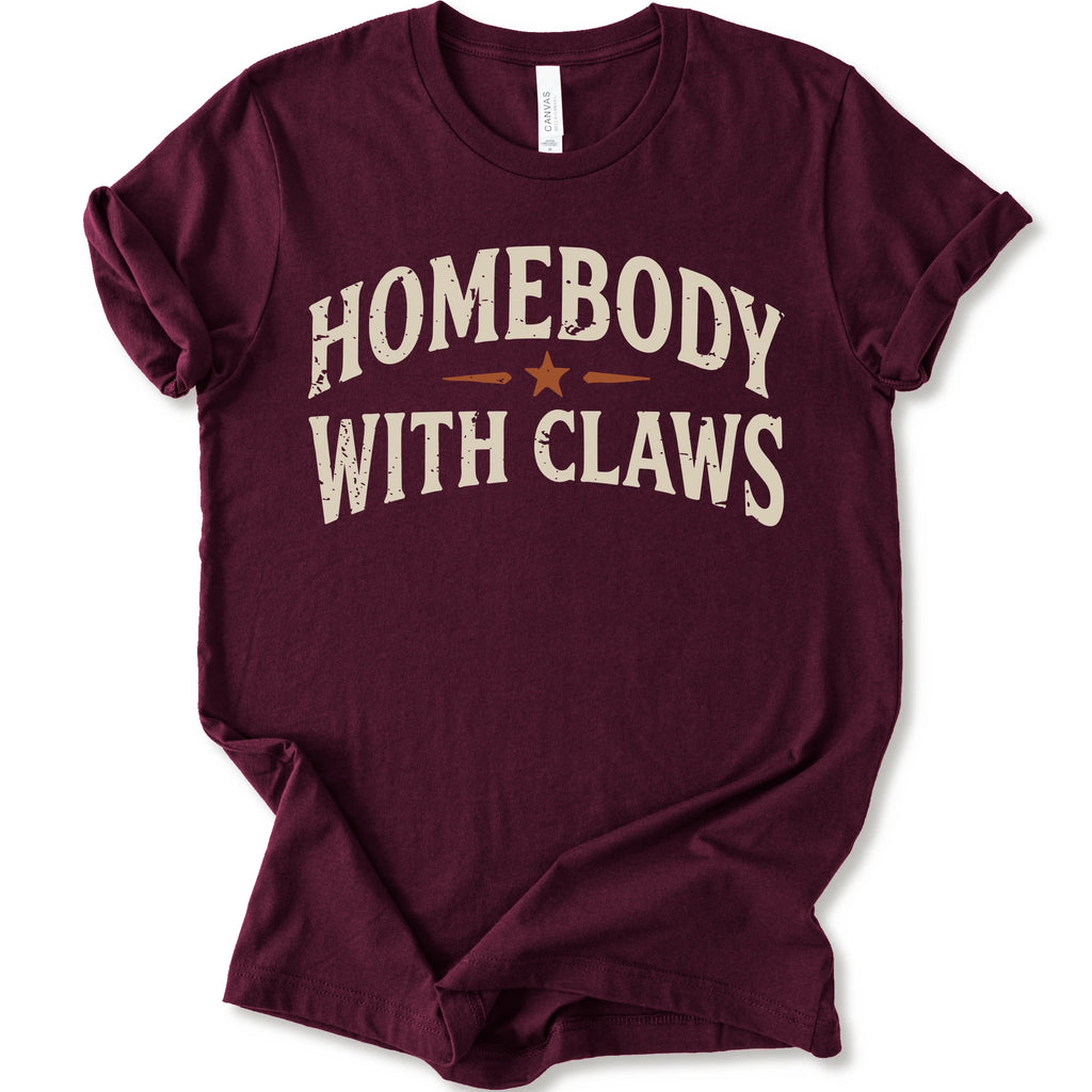 A maroon short-sleeve t-shirt with a distressed graphic reading 'HOMEBODY WITH CLAWS' in cream lettering and a small orange star accent